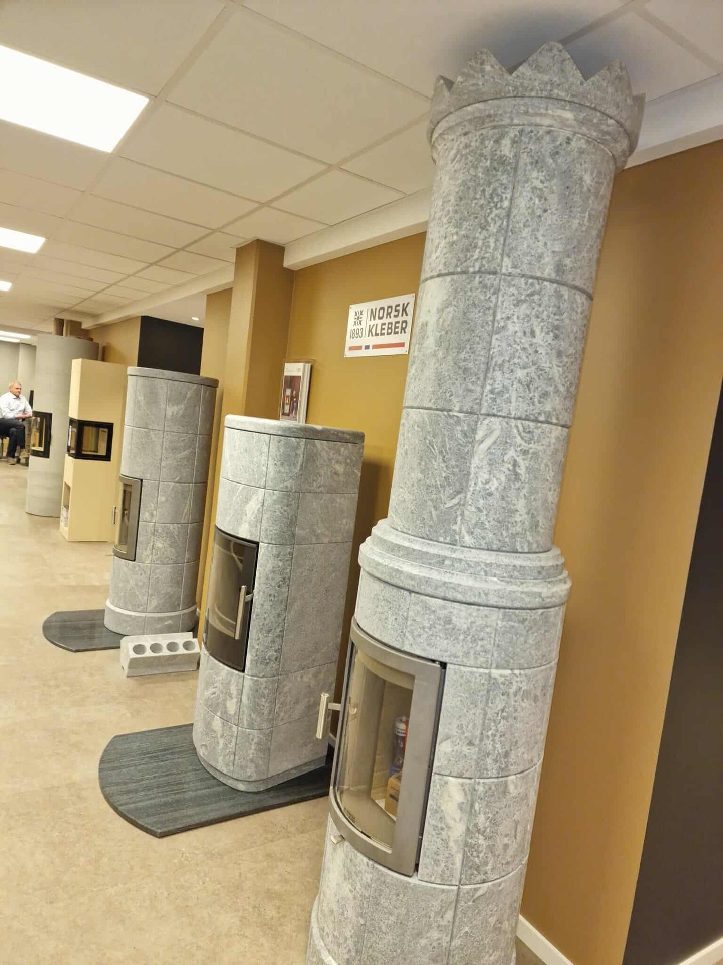 Multiple tall, ornate stone stoves, some with crown tops, displayed in a NORSK KLEBER showroom.