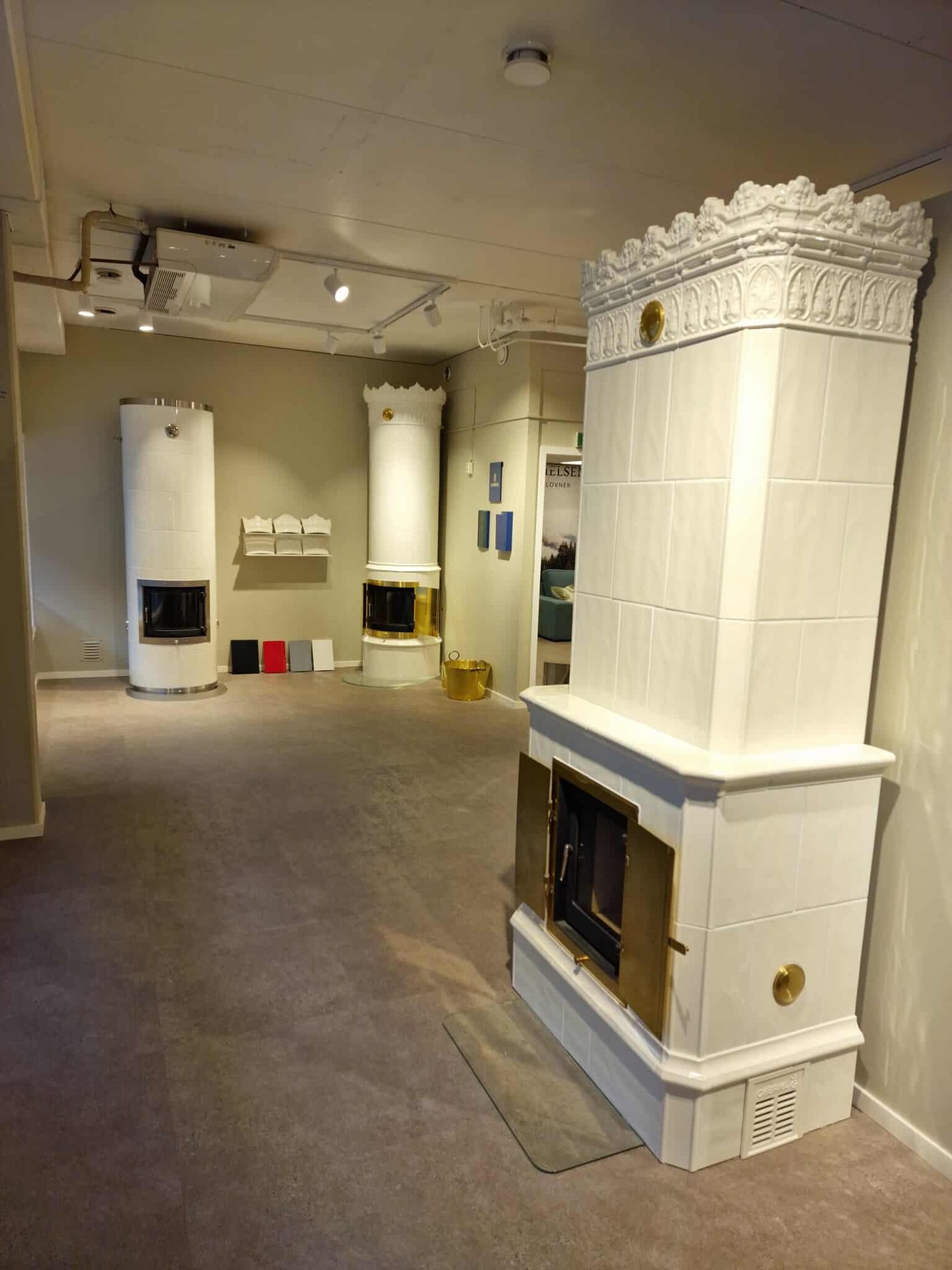 Showroom with three white tiled stoves: one ornate, rectangular with brass doors, two cylindrical.