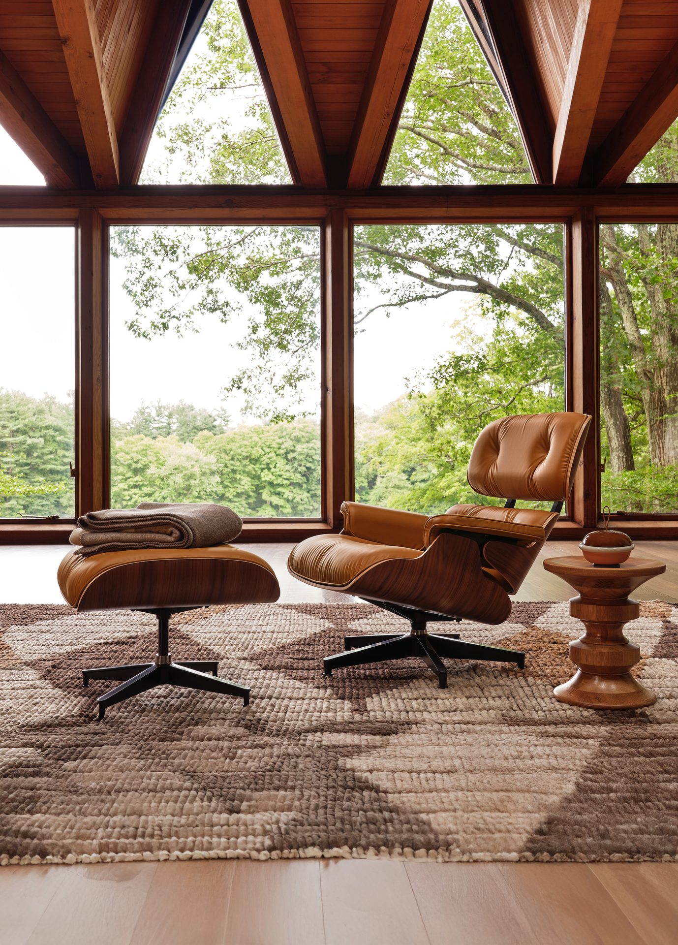 Tan leather chair & ottoman by a large window overlooking a forest, with wood beams.