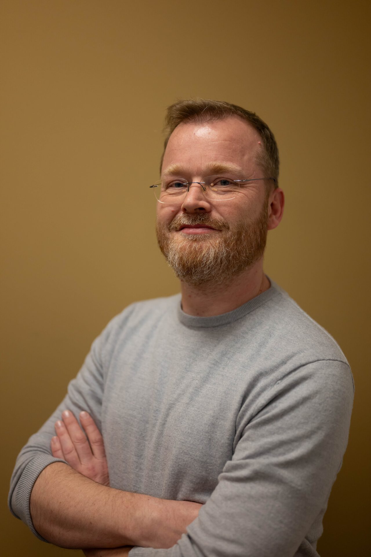 A smiling man with glasses and a beard, wearing a grey sweater, looking directly at the camera.