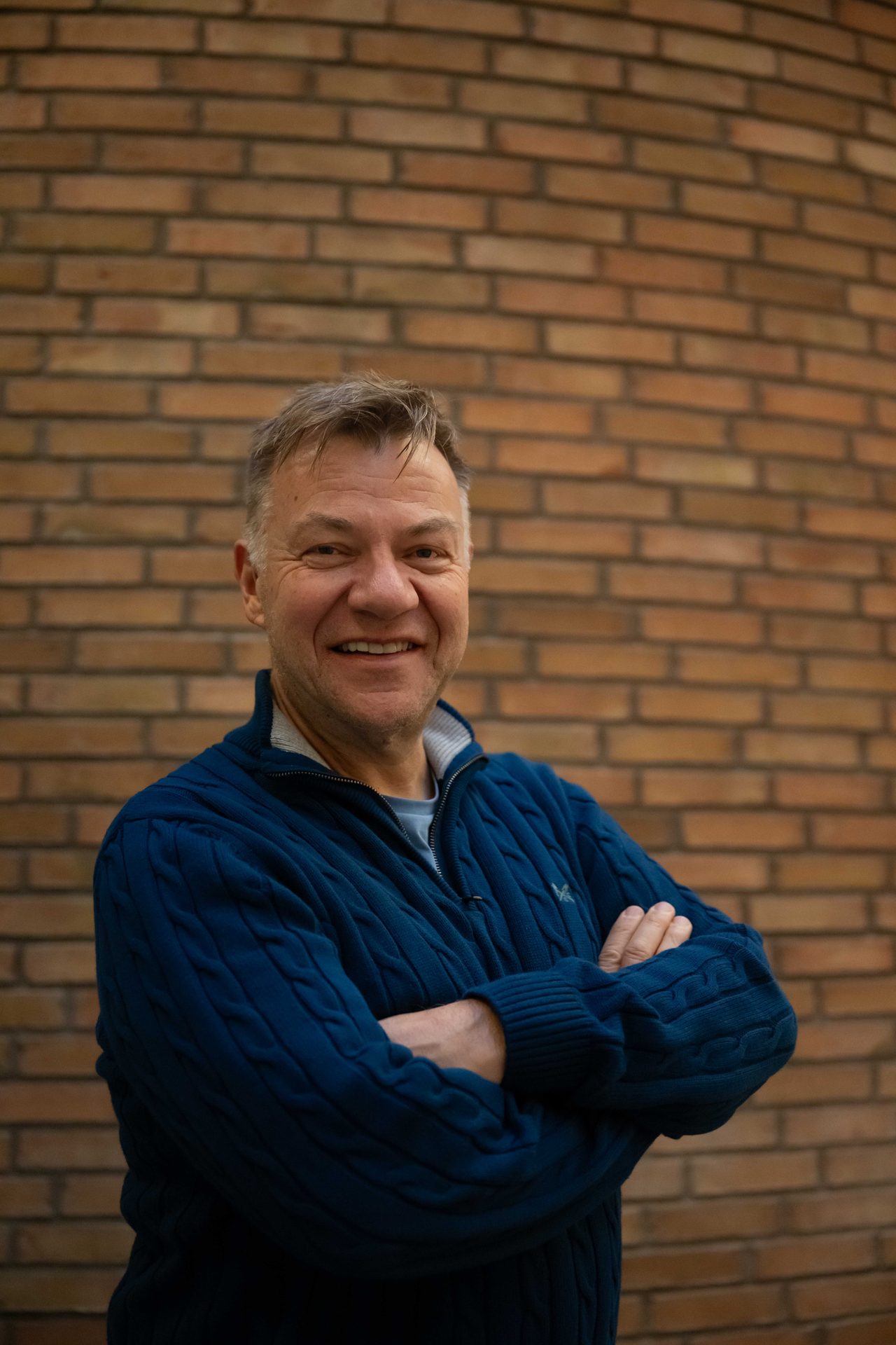A smiling man in a blue sweater stands in front of a brick wall with his arms crossed.