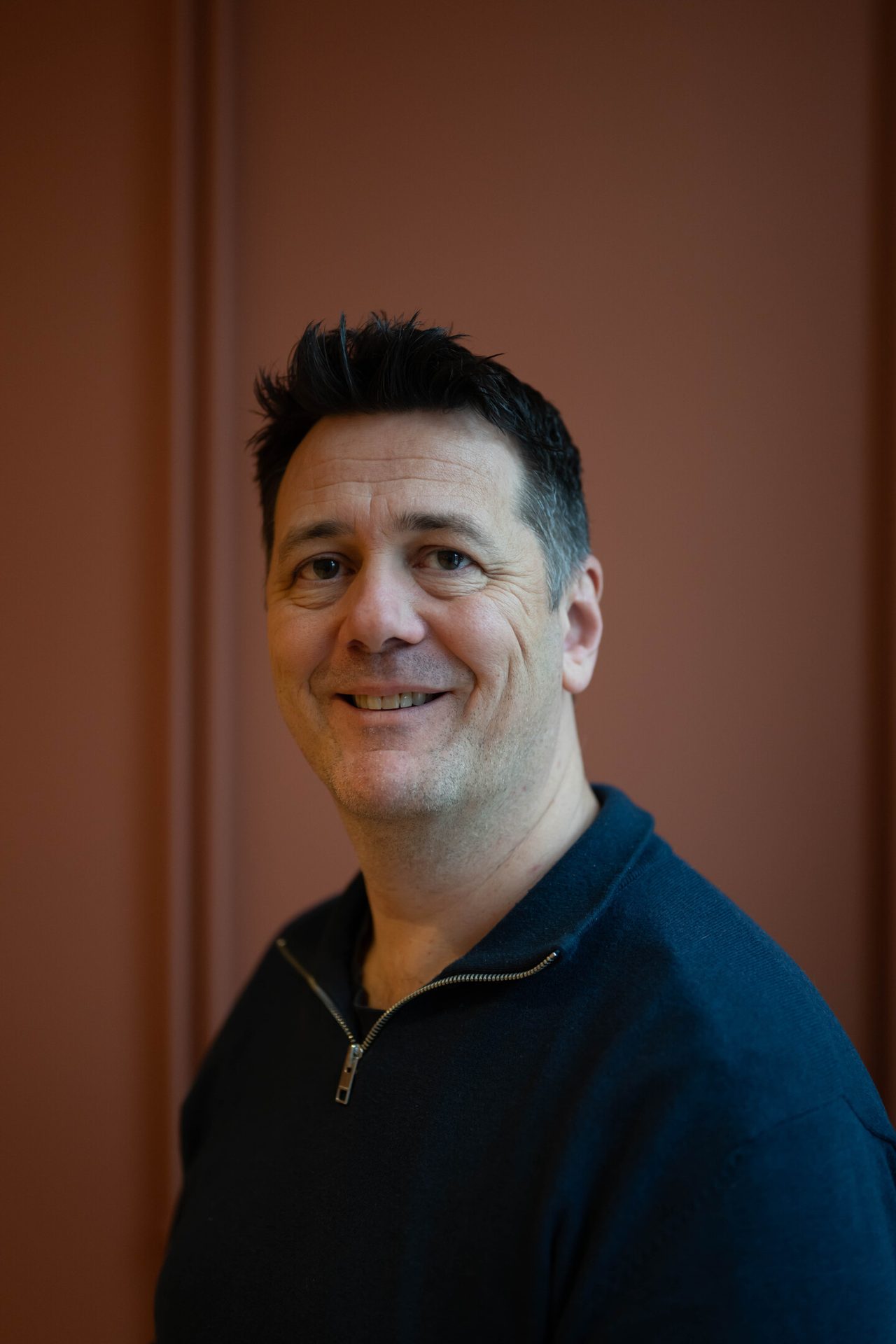 A smiling man in a dark blue sweater, looking directly at the camera with a friendly expression.