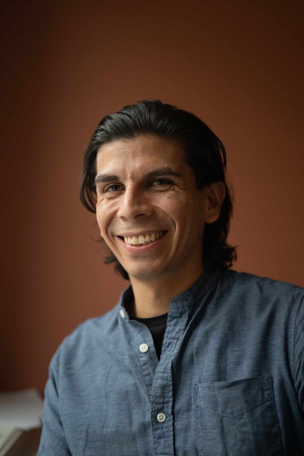 A smiling man with long dark hair and a blue shirt looks directly at the camera.
