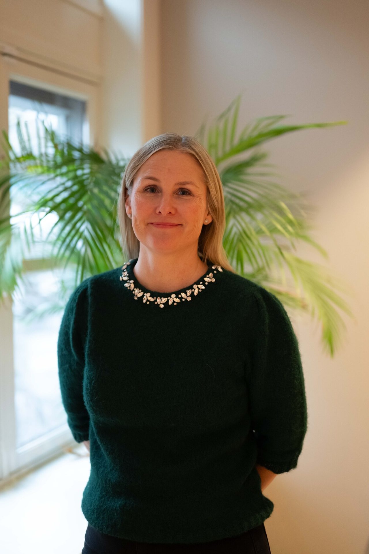Smiling woman in dark green sweater with sparkling embellished neckline, standing indoors.