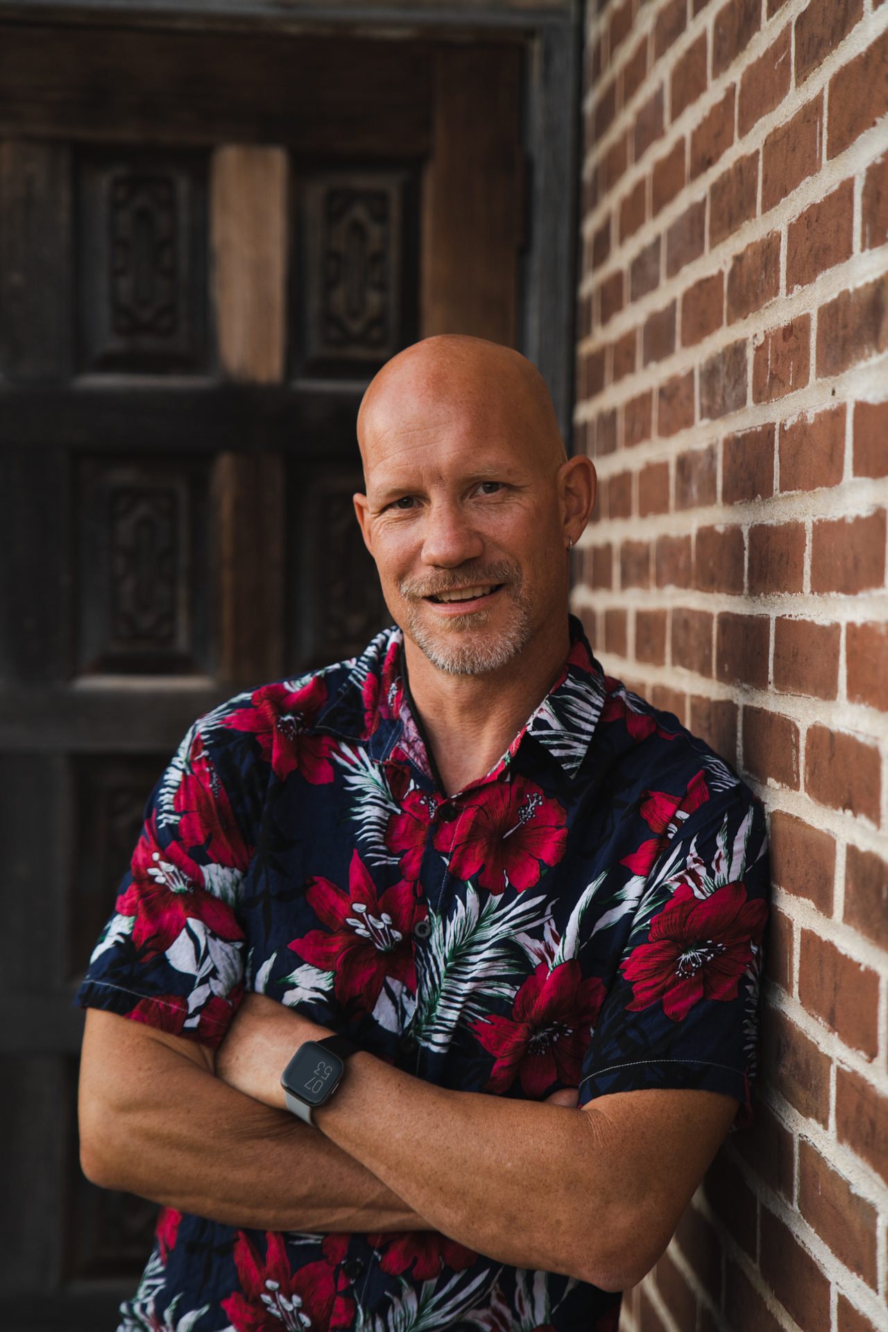 Bald man in a floral shirt with arms crossed, smiling and leaning against a brick wall.