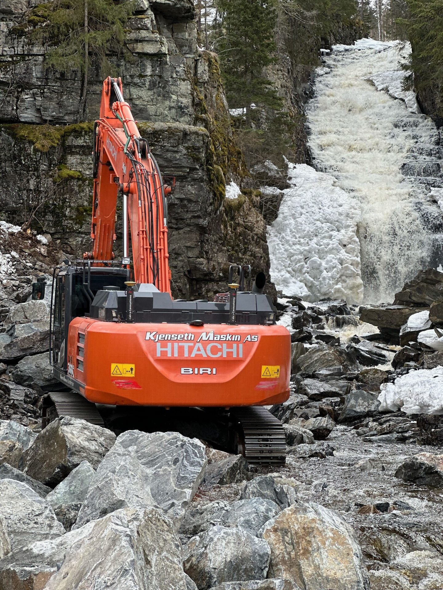 Orange Hitachi excavator on rocks by a powerful icy waterfall.