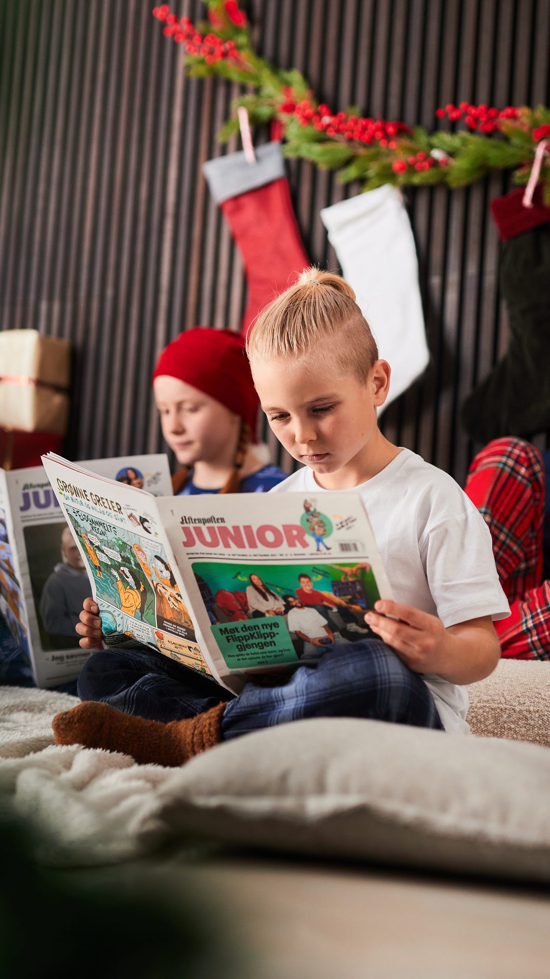 Two young children in pajamas read newspapers in a festive, cozy room.