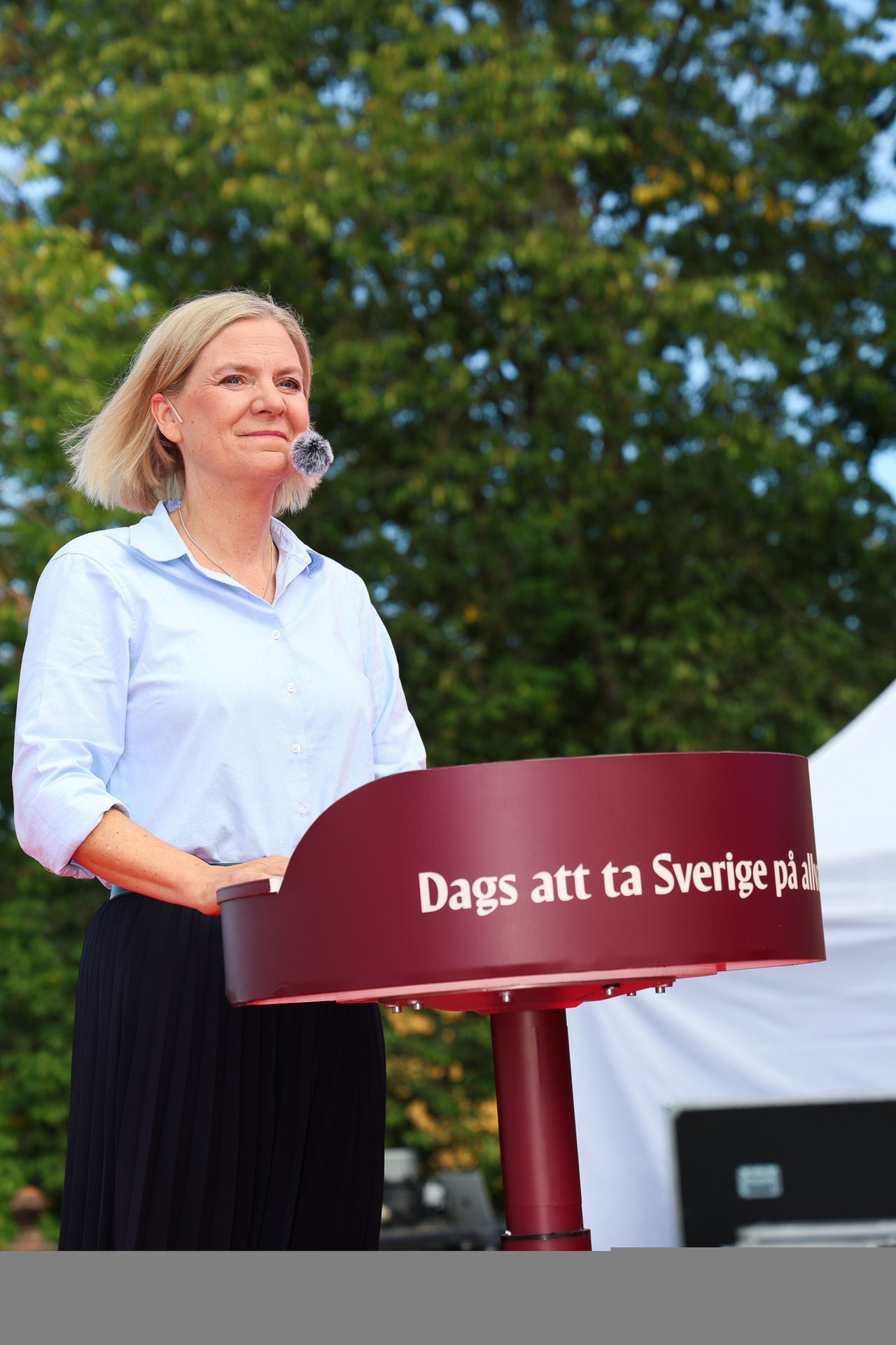 Magdalena Andersson speaking outdoors behind a maroon podium with Swedish text, wearing a light blue shirt.