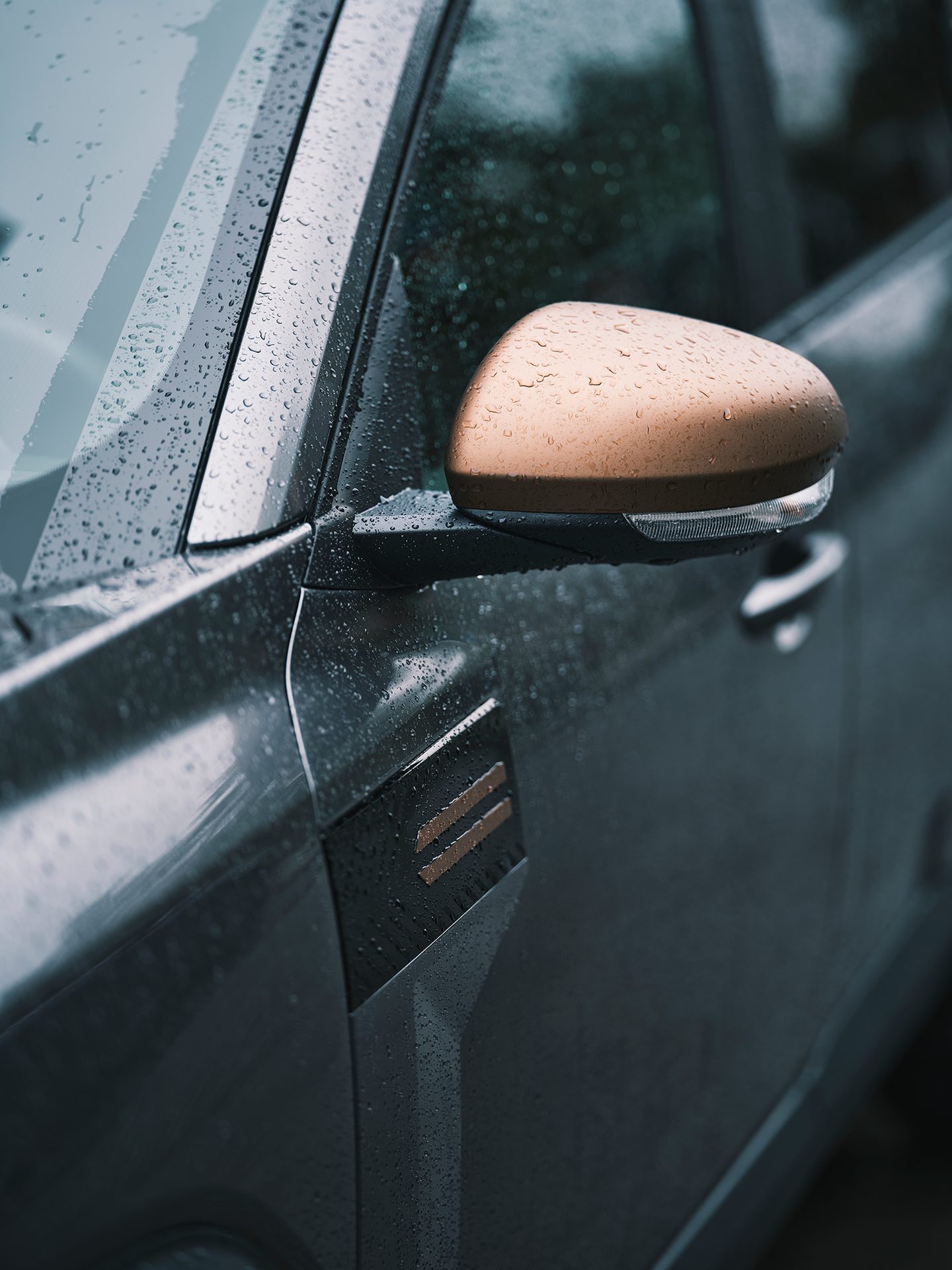 Close-up of a dark car's wet side mirror and body with a bronze emblem, covered in raindrops.