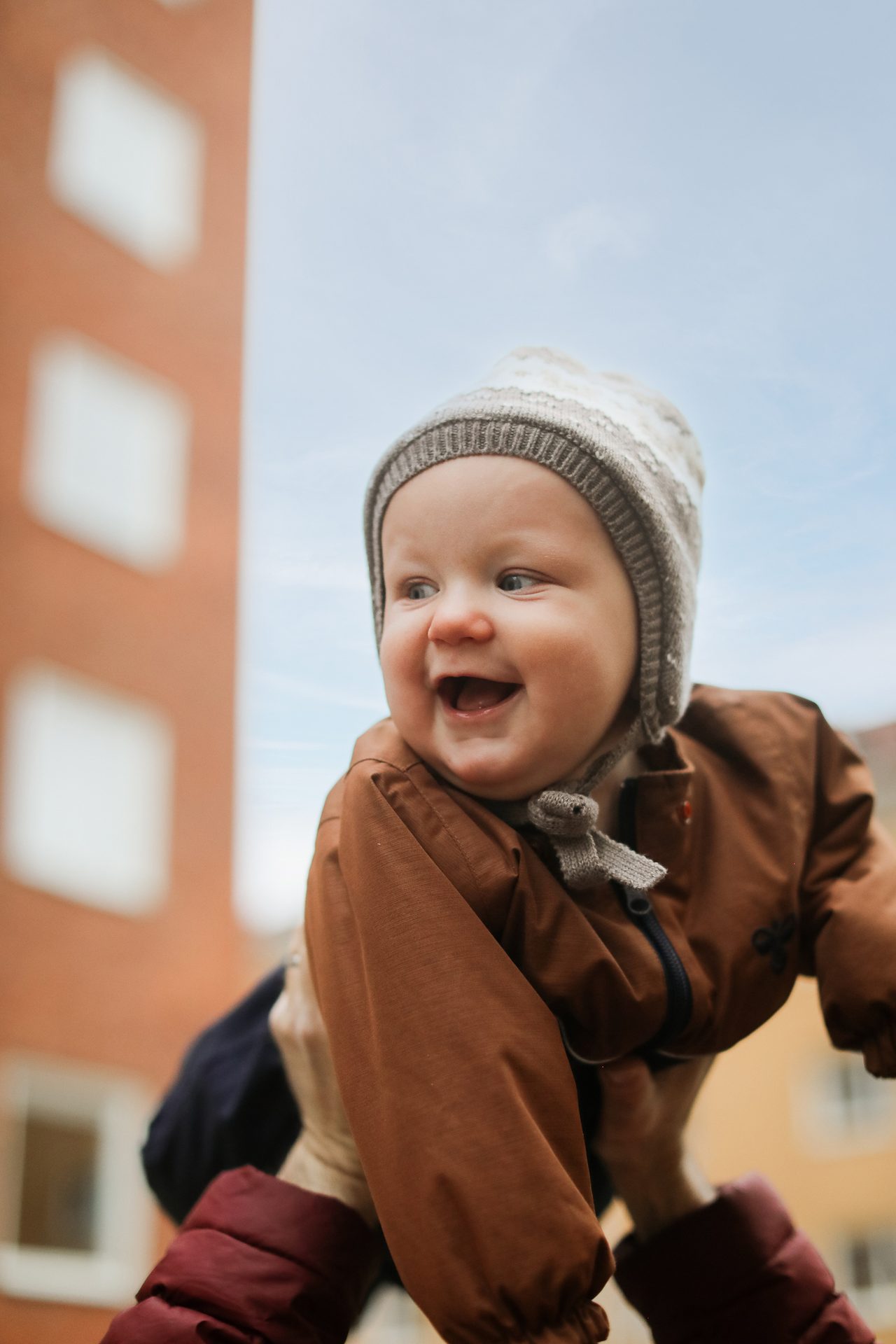 Joyful baby in knit hat and brown jacket, held high, smiling against a building and sky.