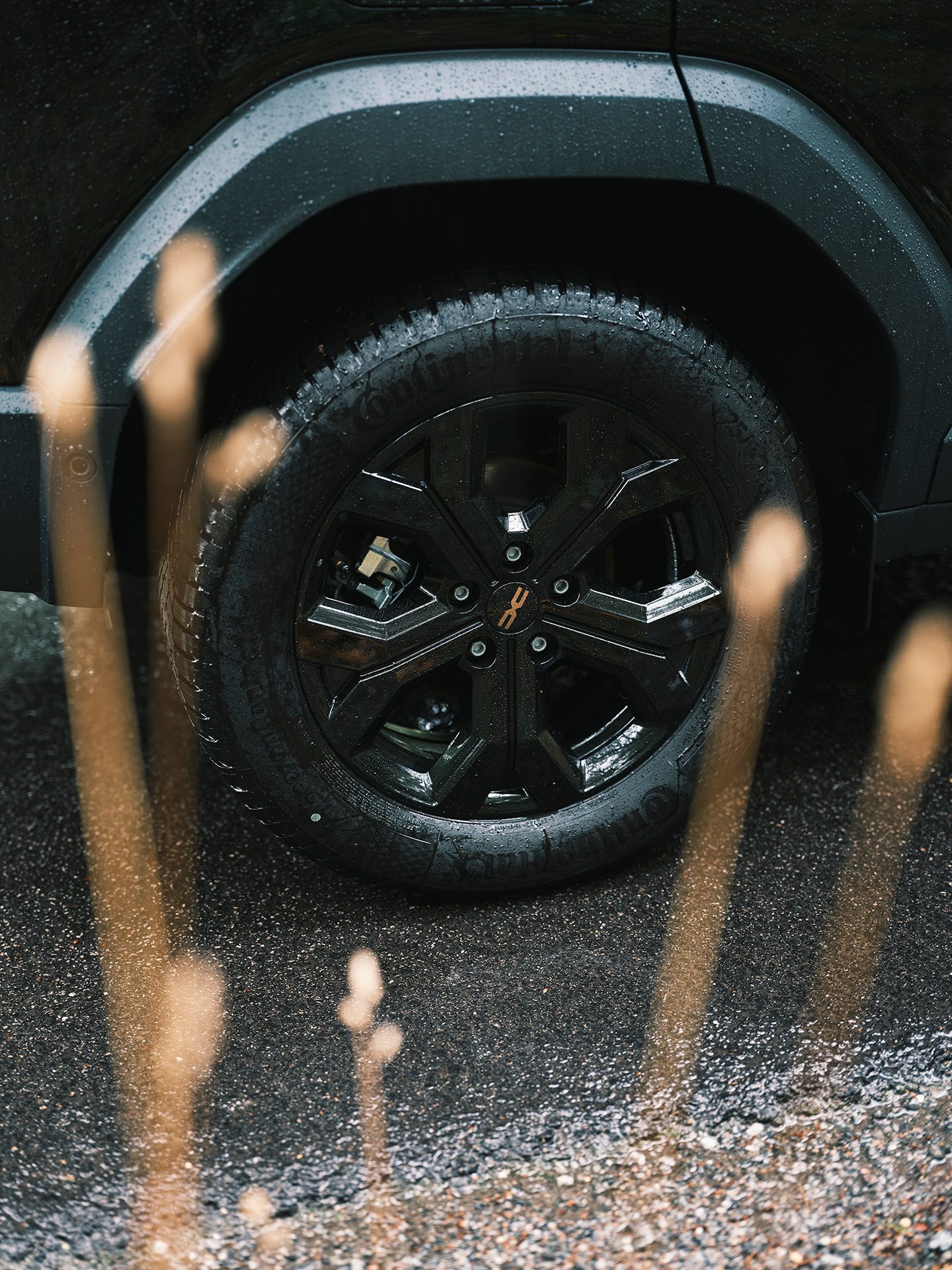 Wet black car wheel, tire, and fender on rainy asphalt, with reflective foreground elements.