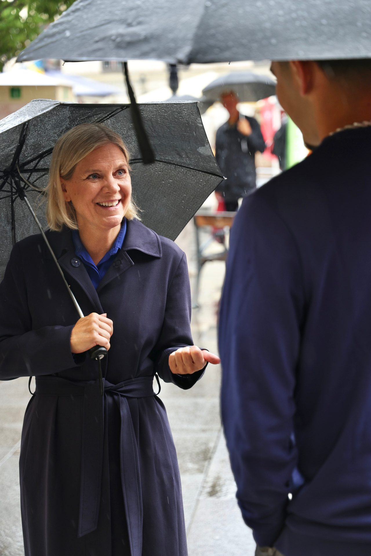 A smiling woman in a navy coat holds an umbrella, gesturing while talking to a man in the rain.
