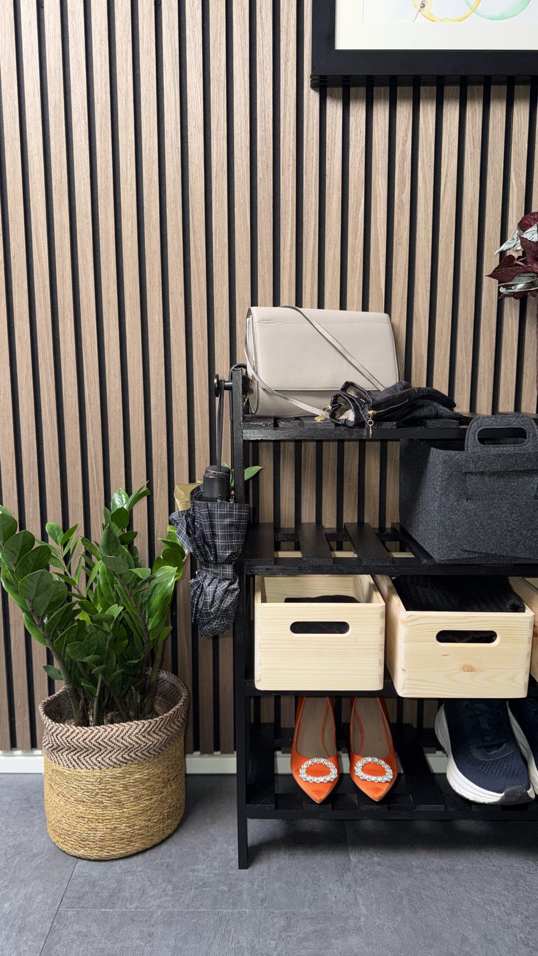 Modern entryway with a wooden slat wall, black shelf holding a plant, handbag, shoes, and storage boxes.