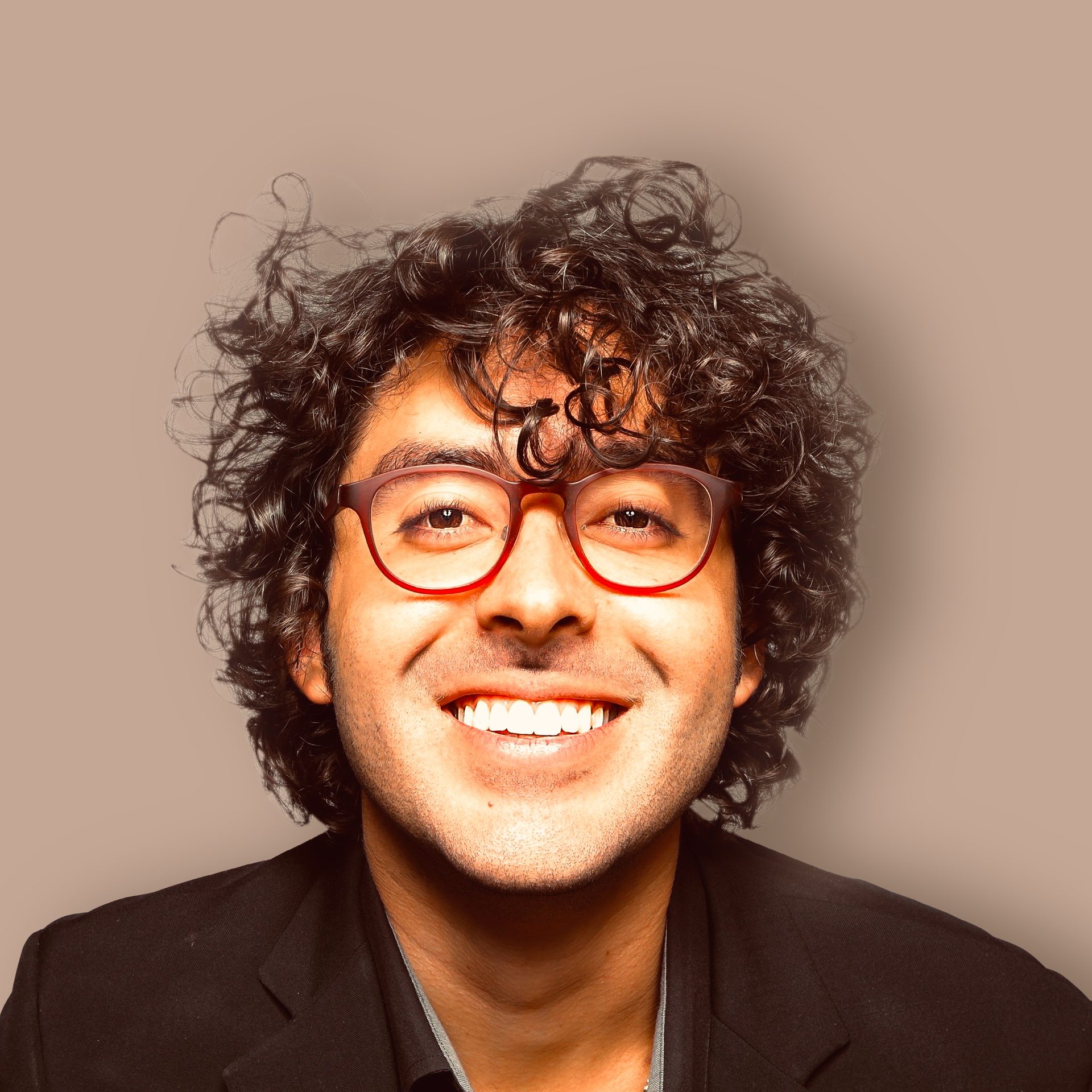 Close-up of a smiling man with curly dark hair and red glasses, wearing a dark jacket.