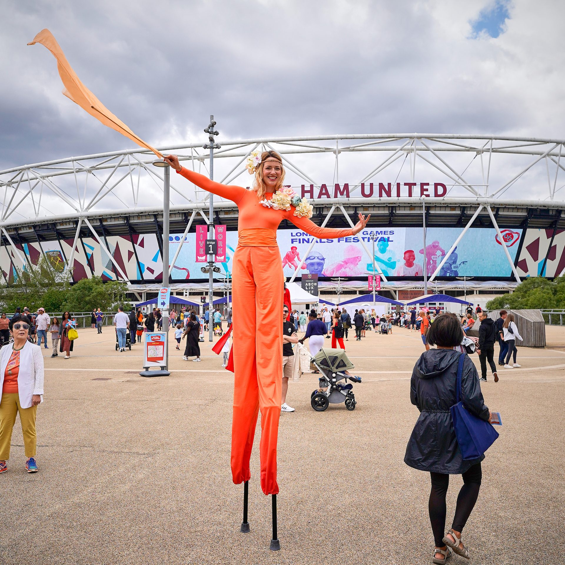 Woman in Orange on stilts, in front of London Stadium
