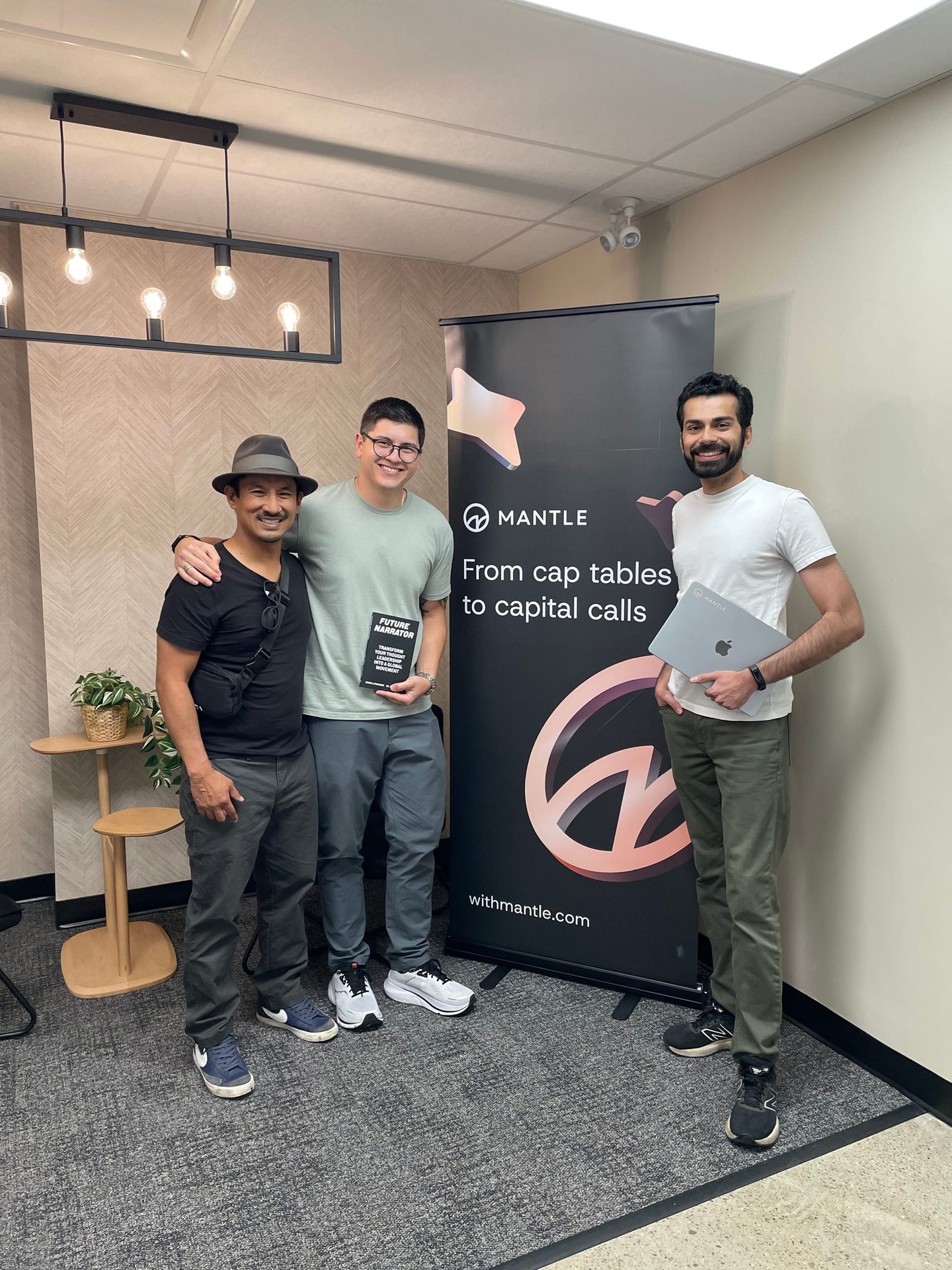 Three men with a 'MANTLE' banner. One holds a 'Future Narrator' book, another a laptop.