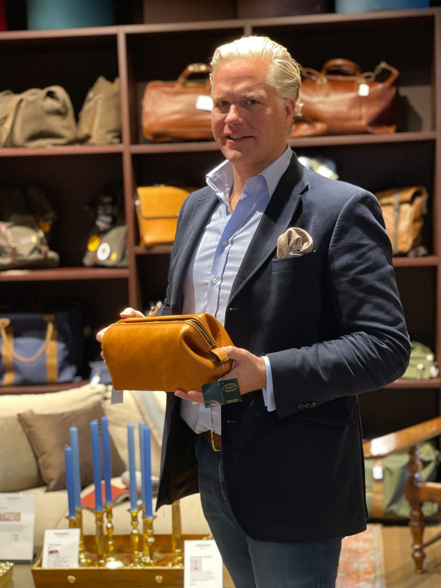A man holds a light brown leather toiletry bag in a store with shelves of other bags.