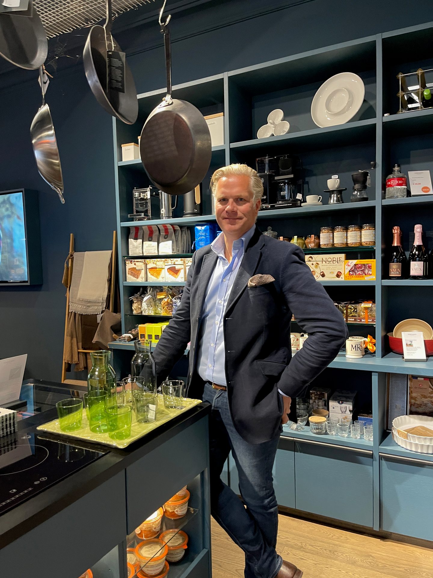 A man in a blazer stands in a kitchenware and gourmet food store, surrounded by shelves and hanging pans.