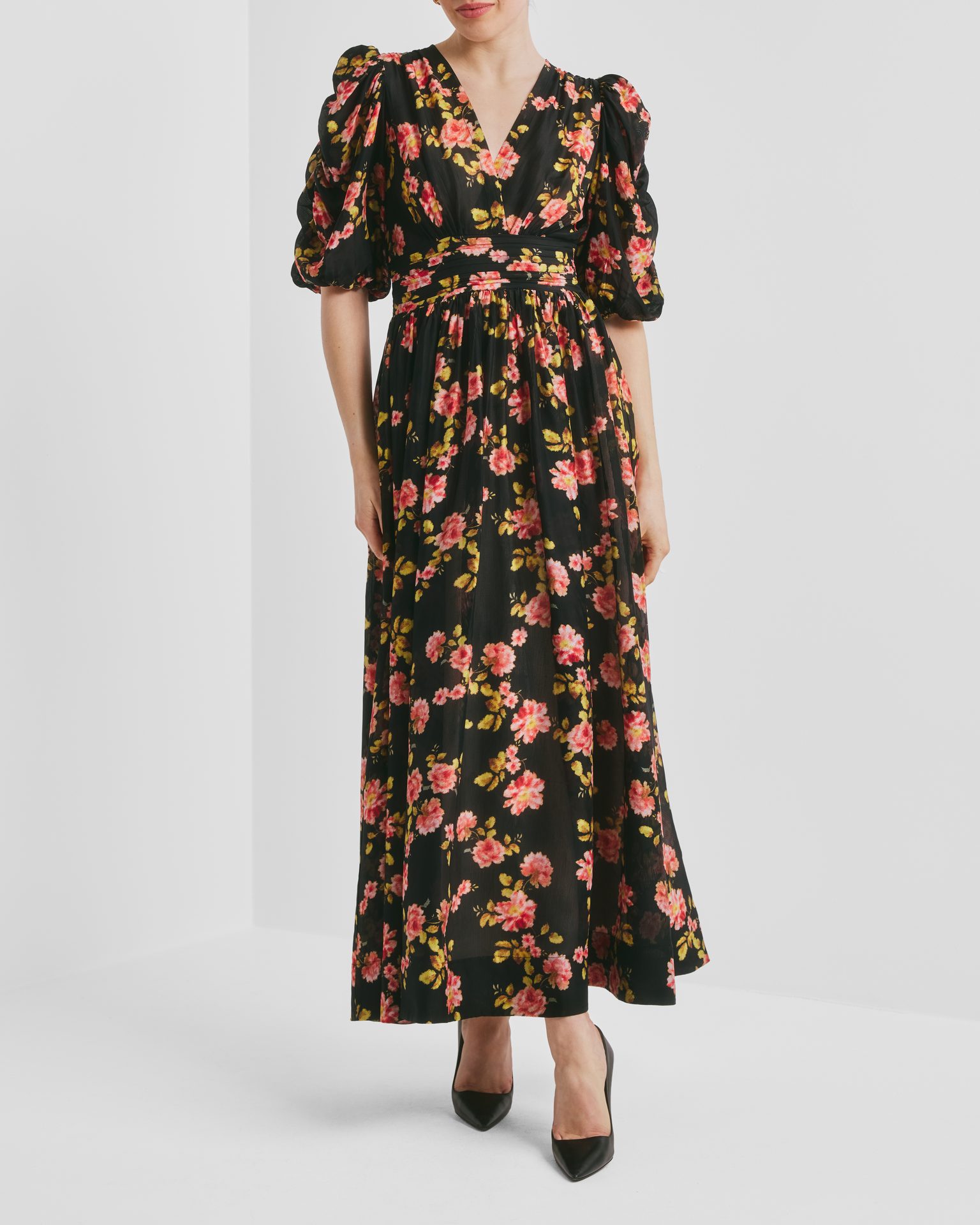 A woman wears a black floral maxi dress with pink and yellow flowers, puffed sleeves, and black heels.