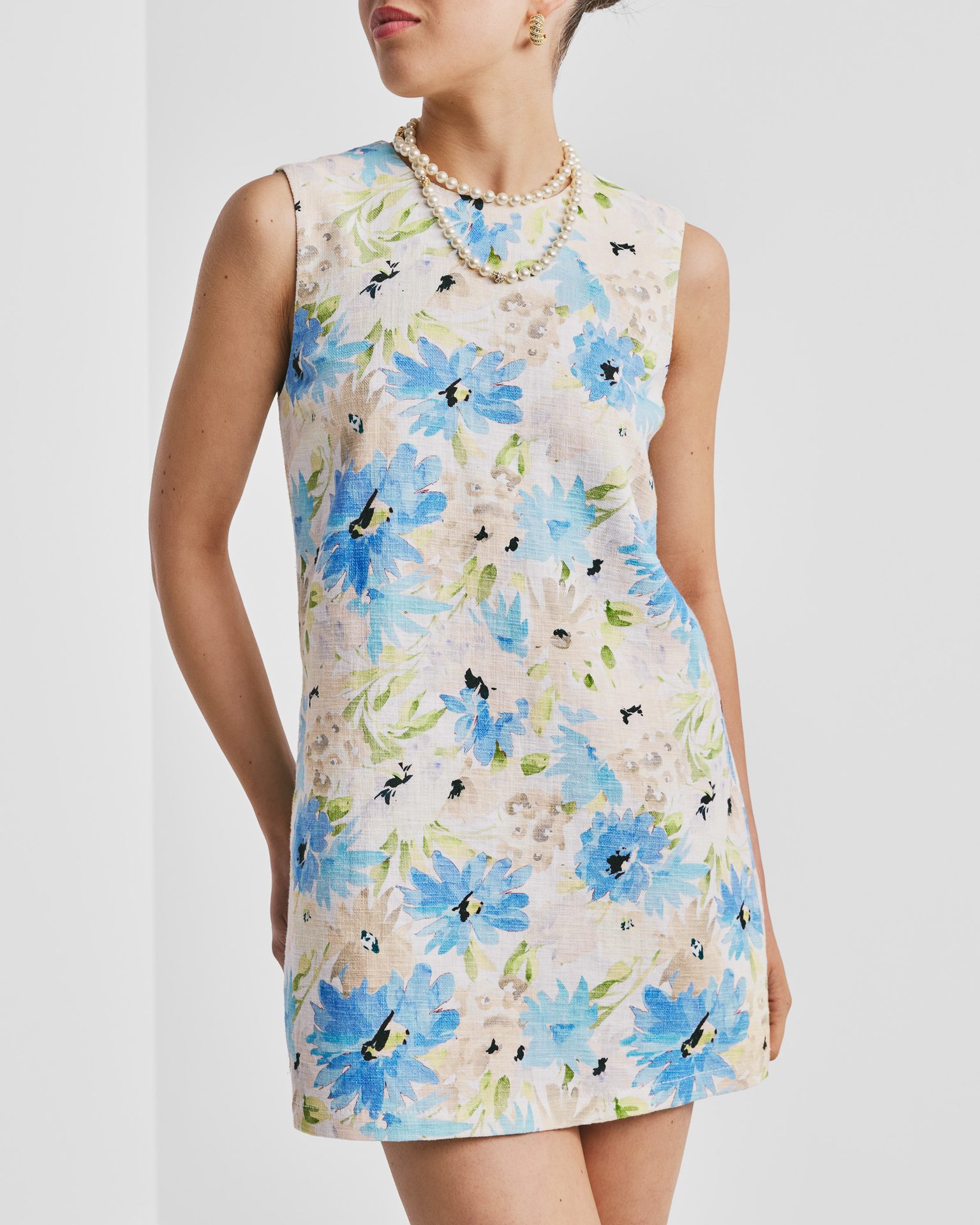 Woman in a sleeveless blue floral print mini dress with a pearl necklace and gold earrings.