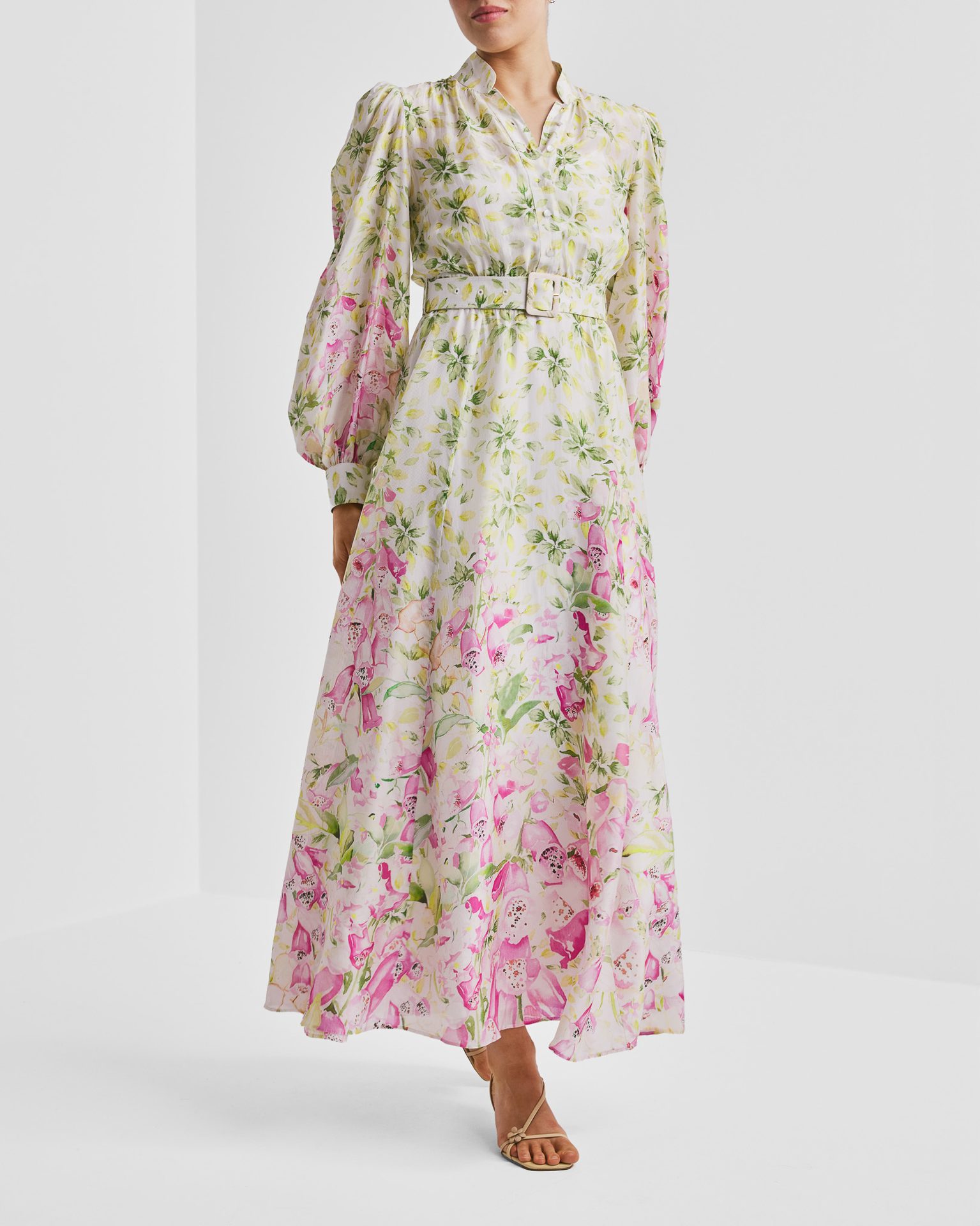 Woman models a white floral maxi dress with pink and green accents, featuring a belted waist and puffed sleeves.