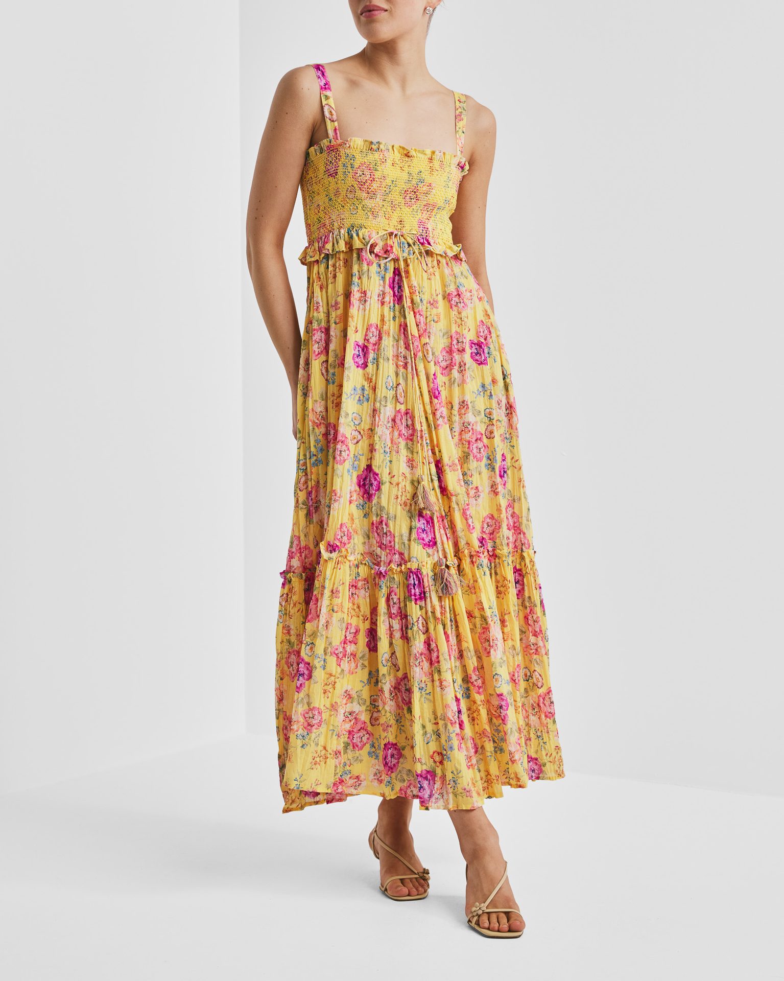 Woman in a vibrant yellow maxi dress with pink, purple, and blue floral print, smocked bodice, and tiered skirt, wearing nude sandals.