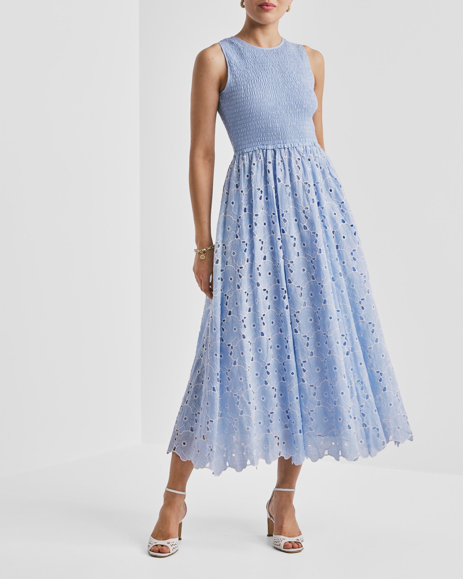 Woman in light blue smocked eyelet midi dress, white heels, and gold bracelet.