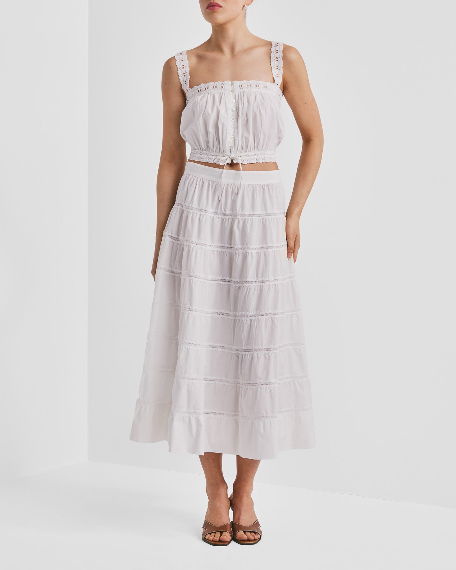 Woman in a white eyelet crop top and matching tiered maxi skirt with brown sandals.
