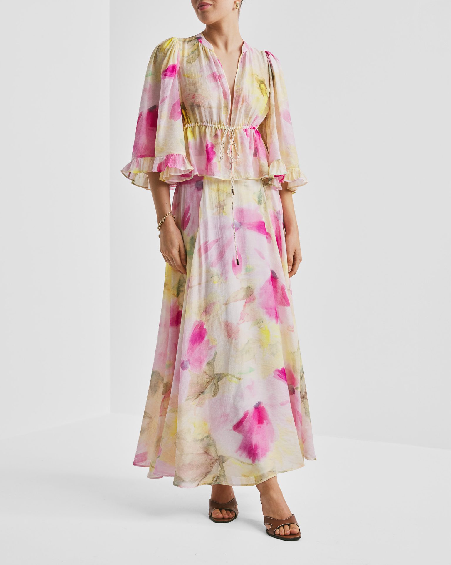 Woman in a long, flowy pink and yellow floral dress with ruffled sleeves, tie waist, and sandals.