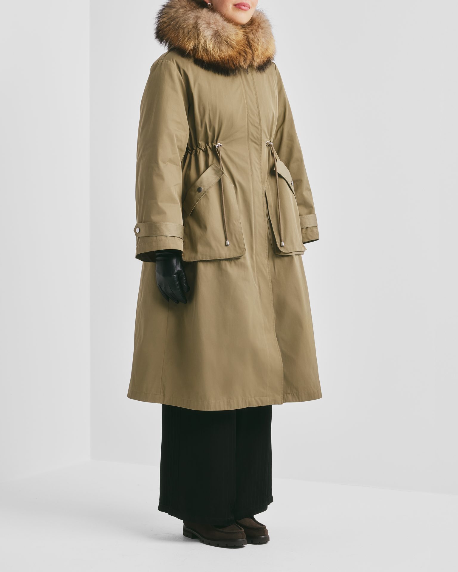 Person in an olive green fur-hooded coat, black gloves, pants, and dark shoes.