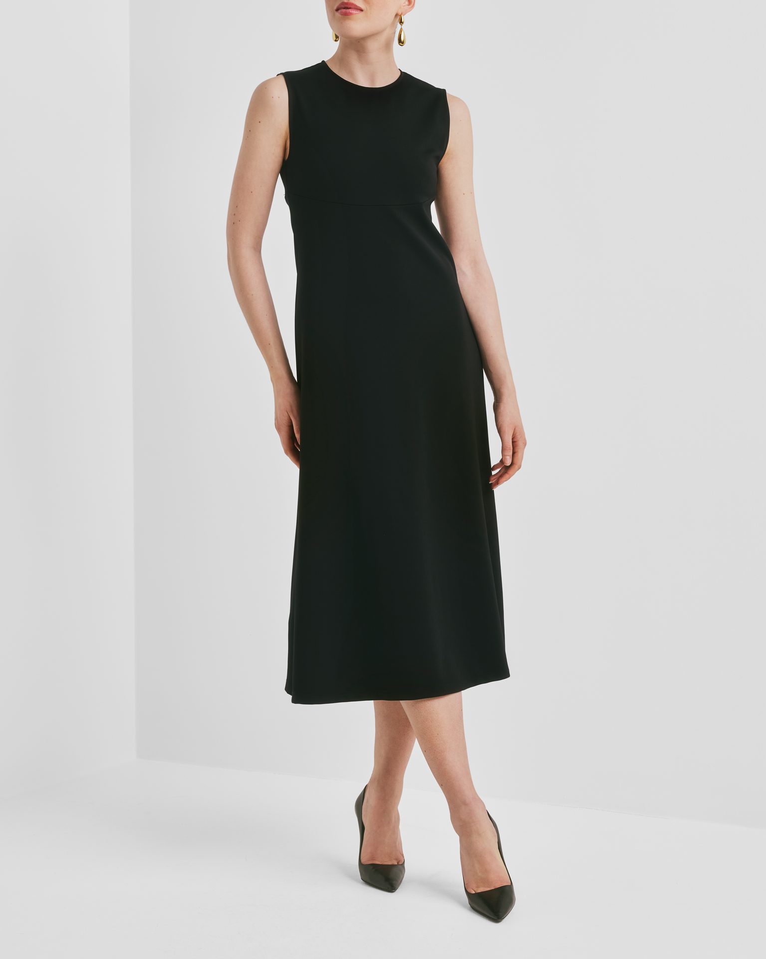 A woman in a black sleeveless midi dress and black heels, posing against a white background.