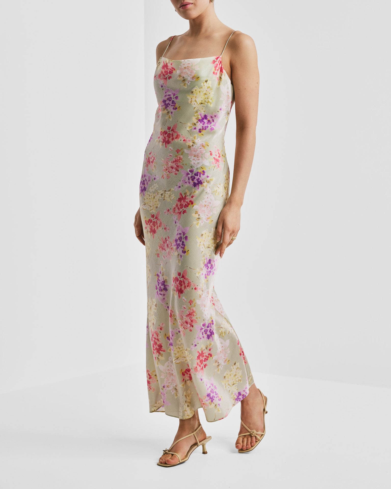 A woman in a light green floral maxi slip dress with pink and purple flowers, wearing beige heels.