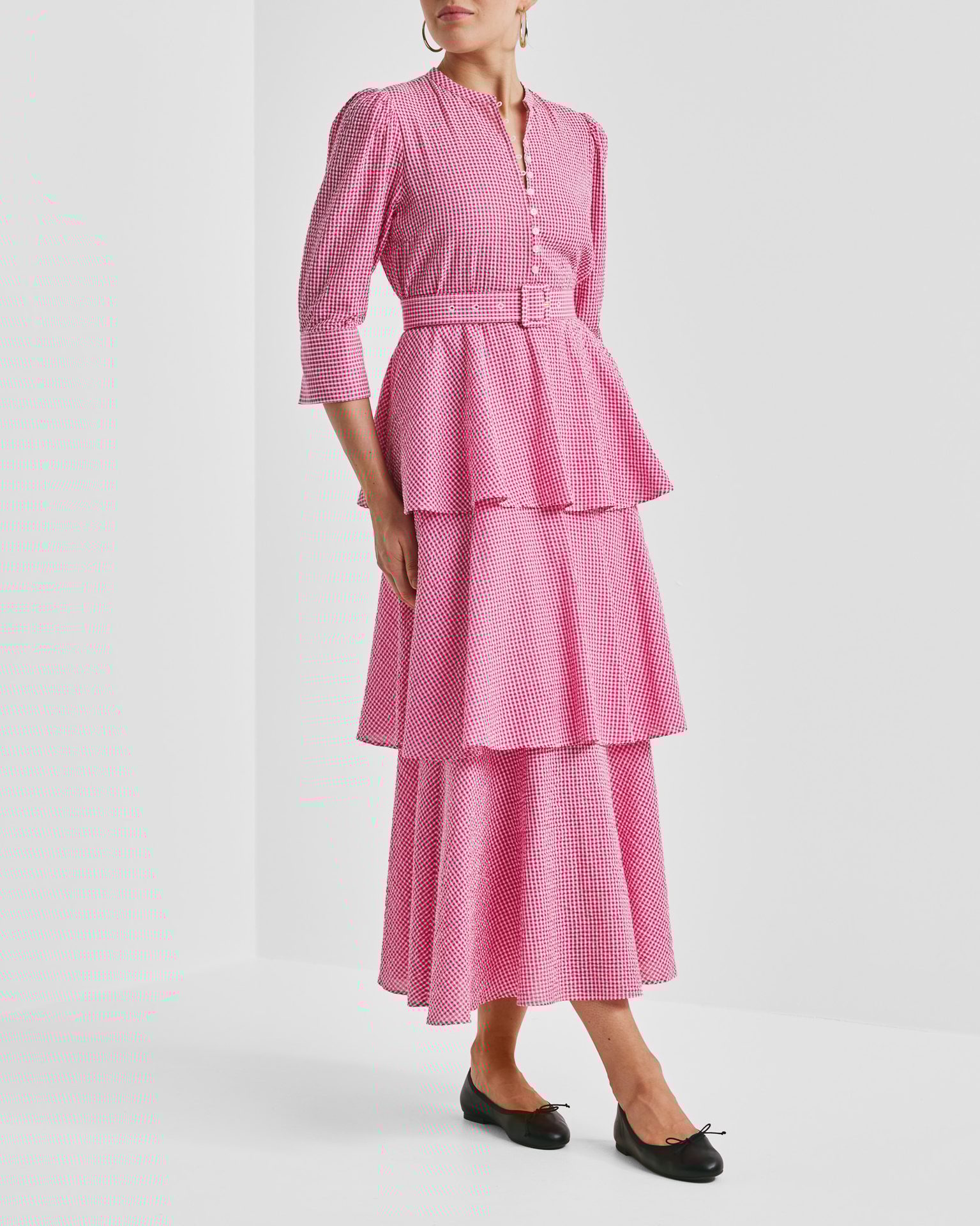 Woman in a pink gingham tiered midi dress with a belt and black ballet flats.