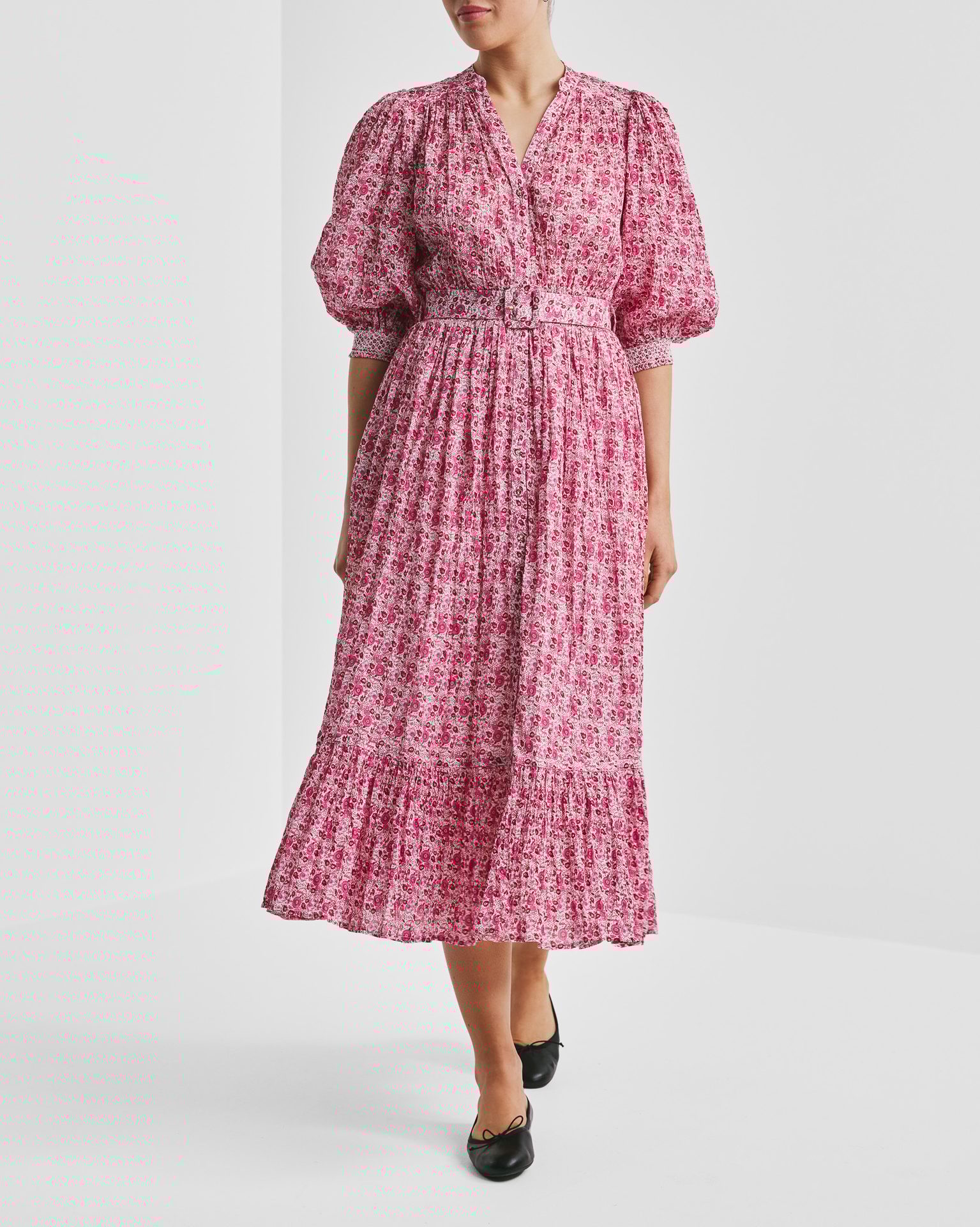 Woman wearing a pink floral midi dress with puff sleeves, a V-neck, belt, and black flats.