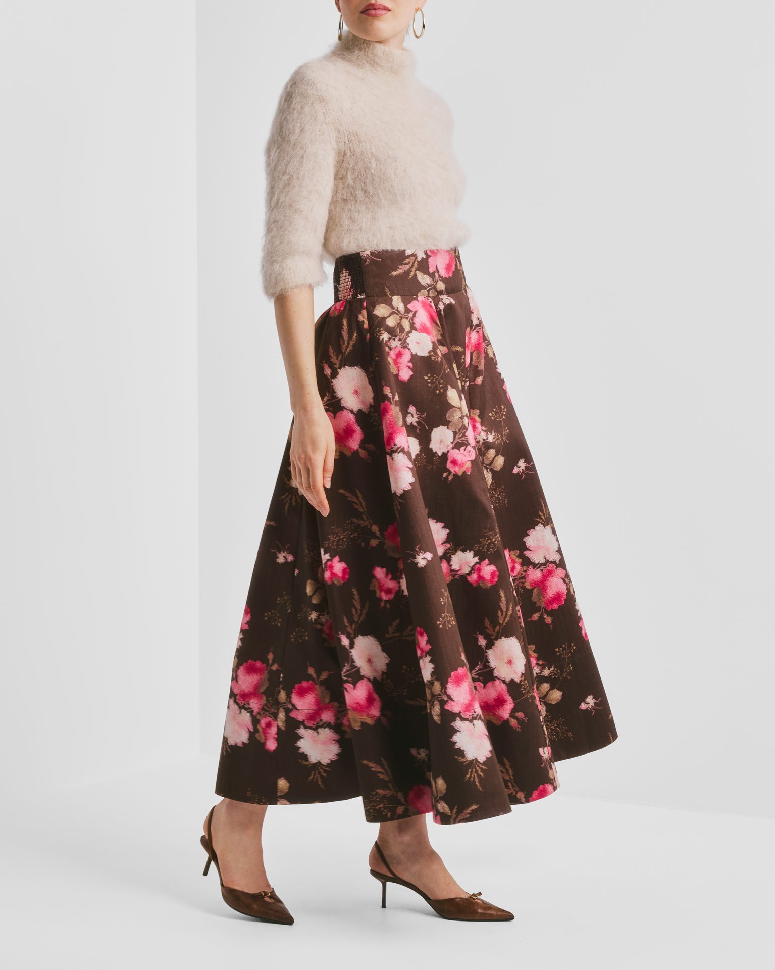 Woman in fuzzy cream sweater, brown floral skirt, and brown heels, standing at an angle.