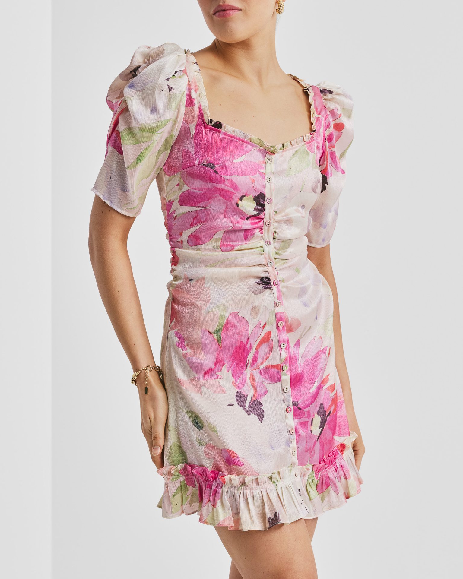 A woman models a short, pink and green floral print dress with puff sleeves, a button-front, and ruffled hem.