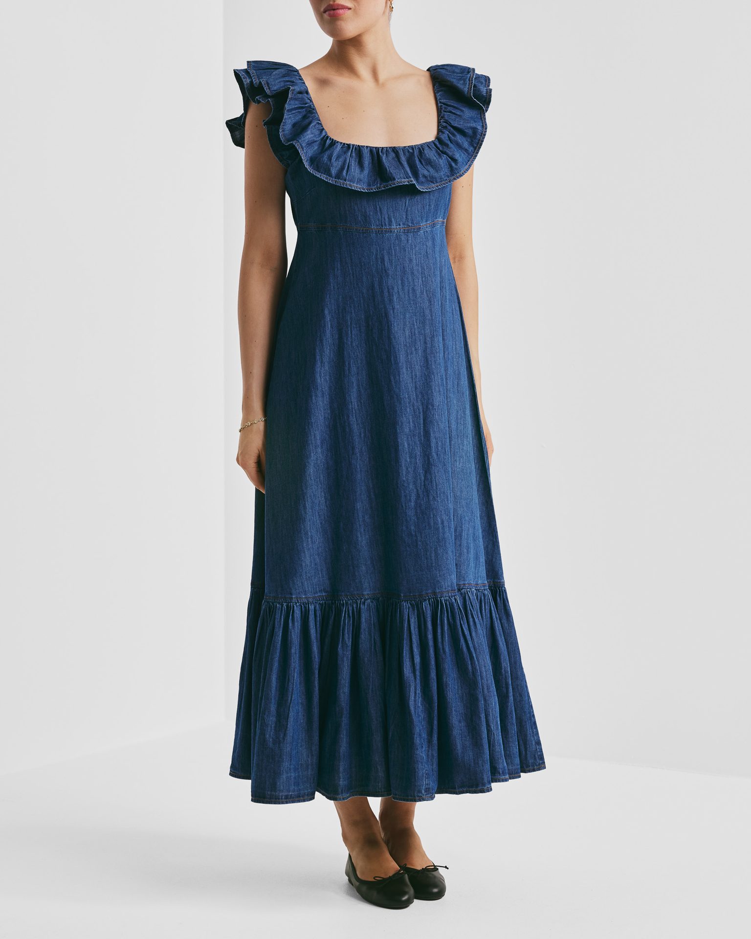 A woman wears a blue denim dress with a ruffled square neckline, ruffled hem, and black flats.