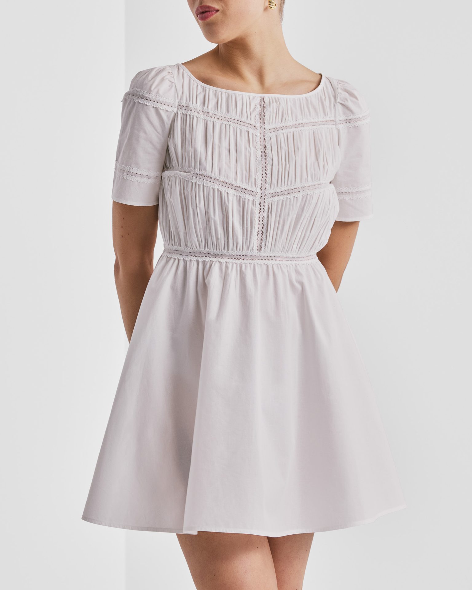 A woman in a white short-sleeve dress with pintuck and lace details on the bodice and a gathered waist.