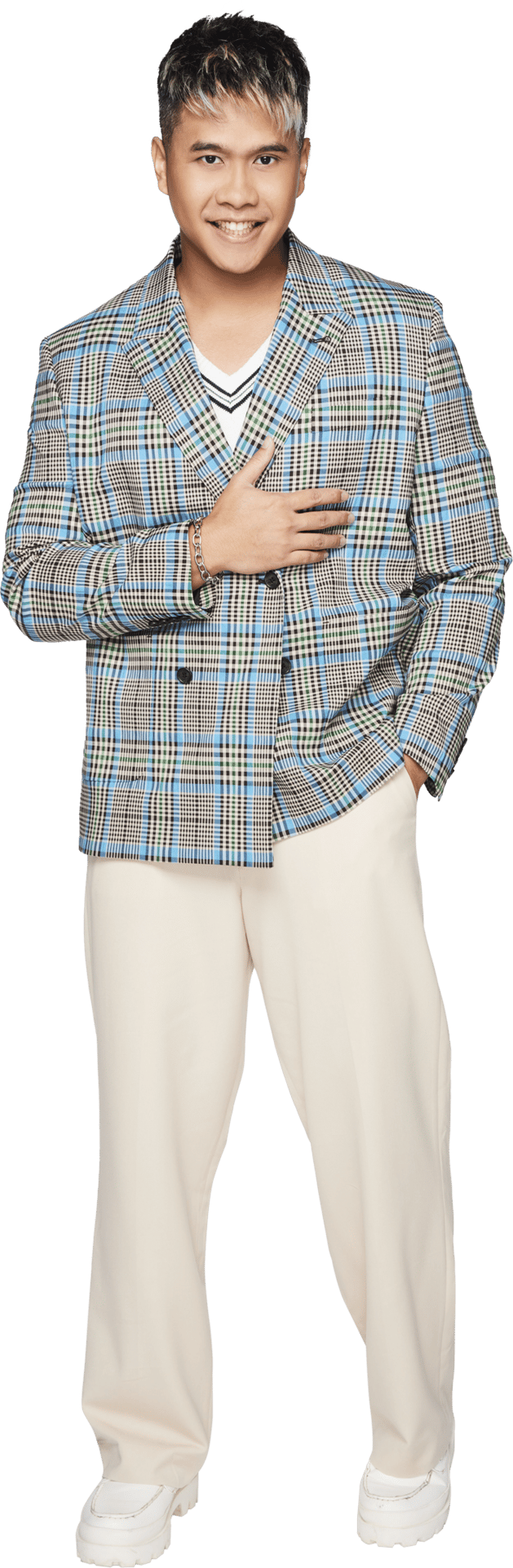 Smiling man in blue plaid blazer, beige pants, white sneakers, and highlighted hair, hand on chest.