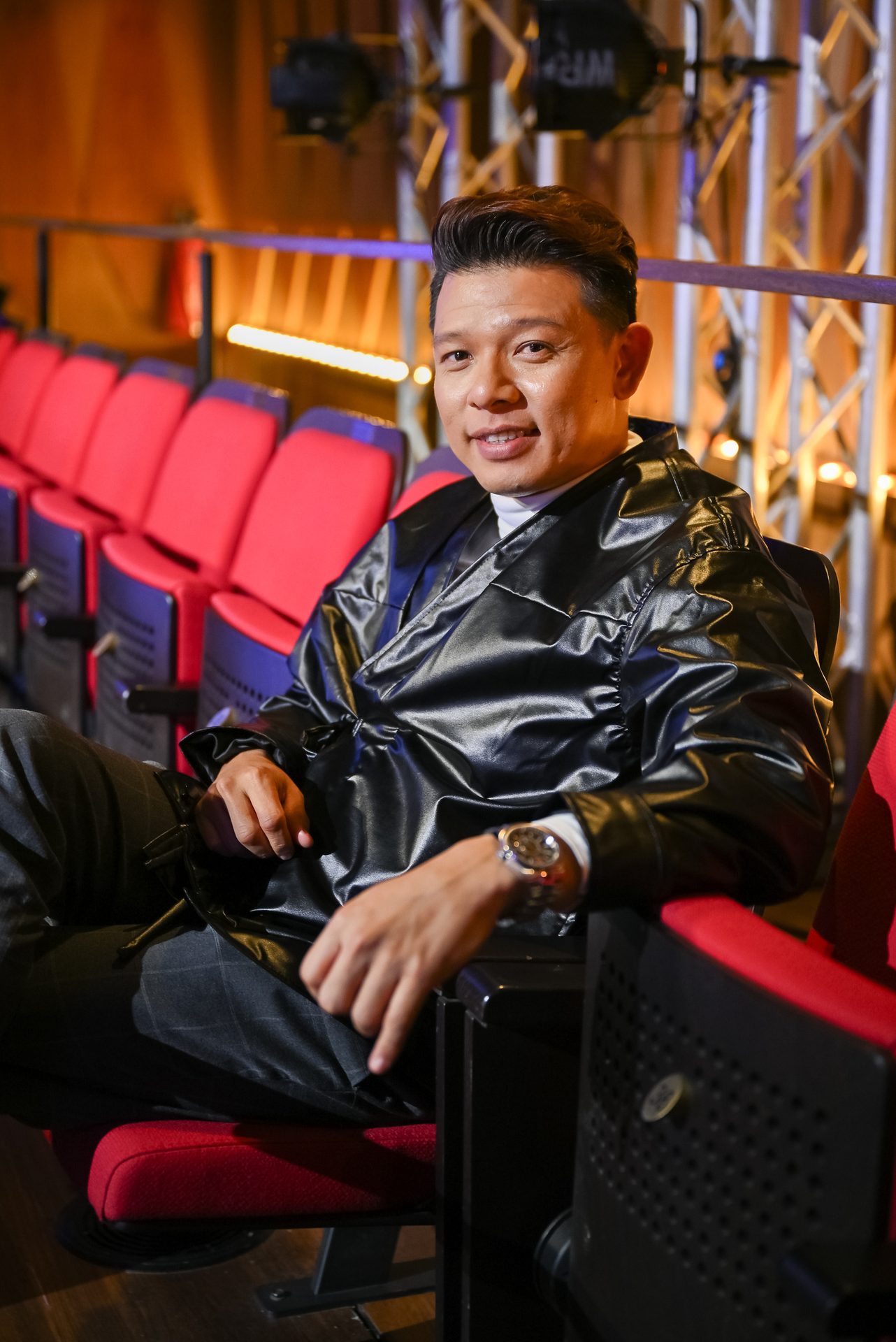 Smiling man in black jacket sits in red auditorium seats.