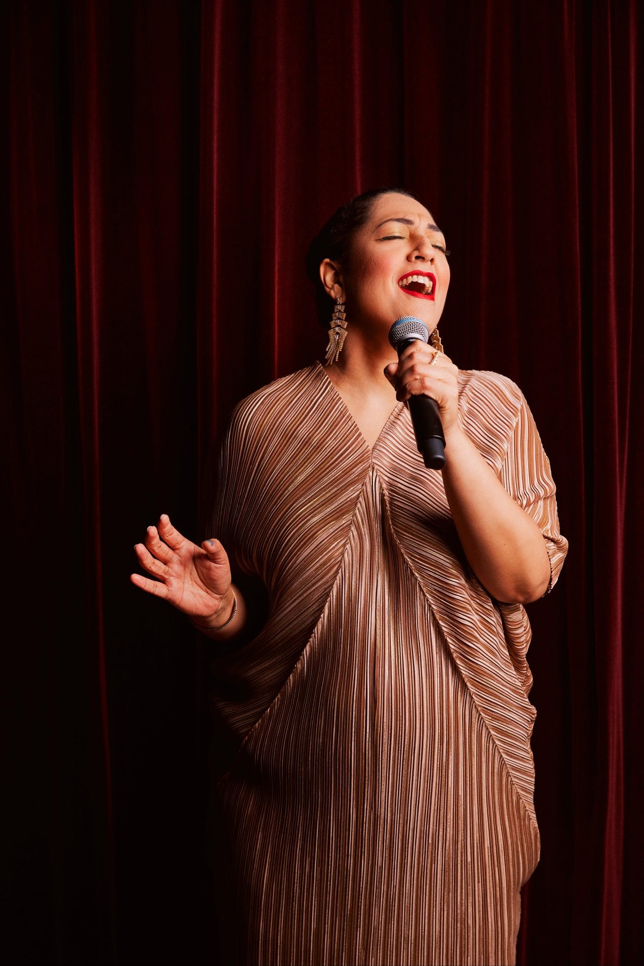 Woman singing into a microphone, eyes closed, wearing a pleated golden outfit against red curtains.