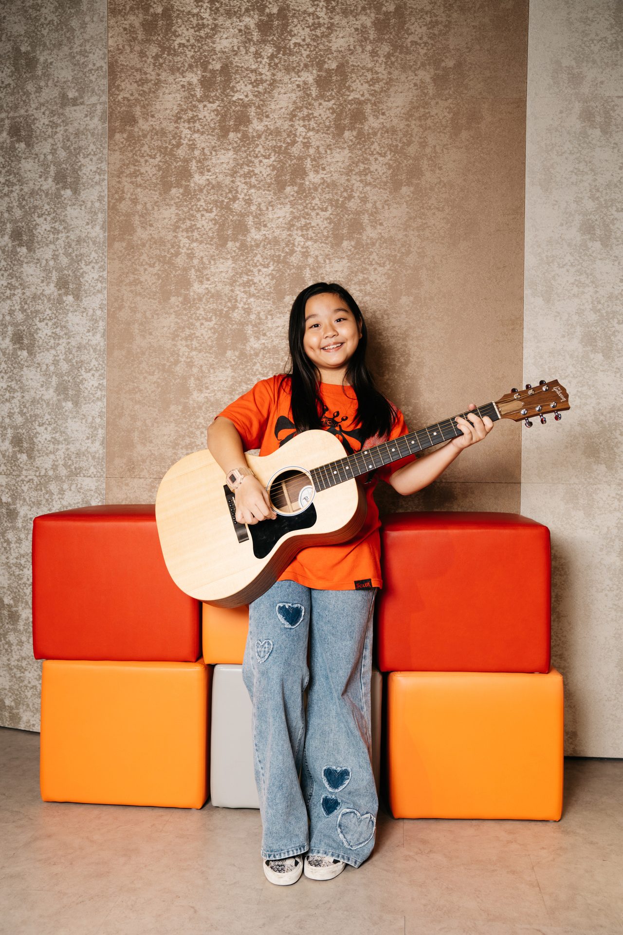 Smiling Asian girl in orange shirt and heart jeans holds acoustic guitar by colorful blocks.