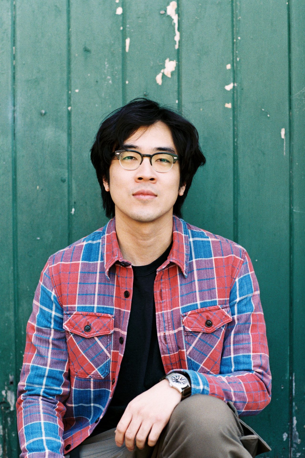Young man with dark hair, glasses, and a red/blue plaid shirt, looking forward against a green wall.