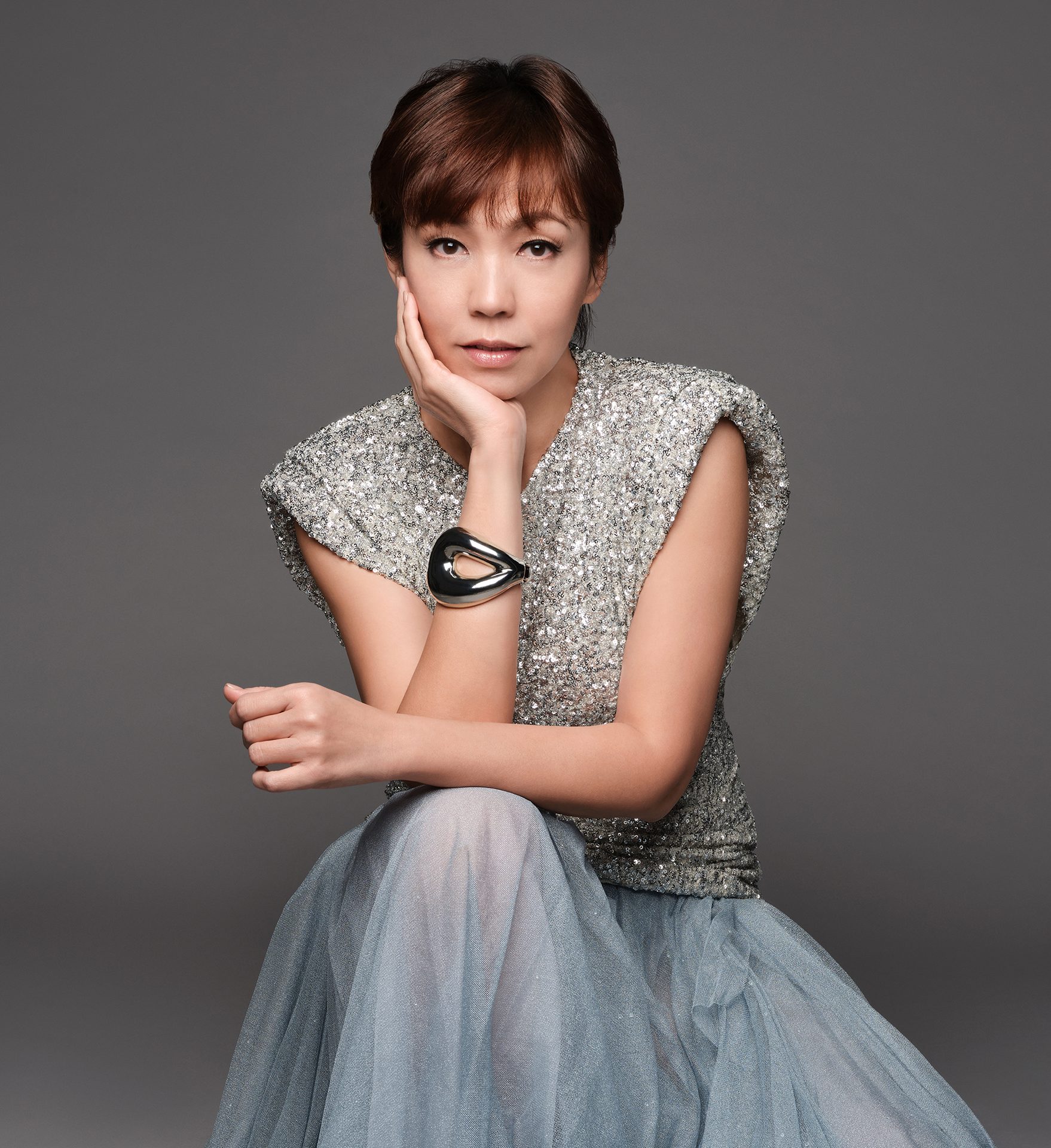 Woman in sparkling top & blue skirt, hand on chin, looking at camera.