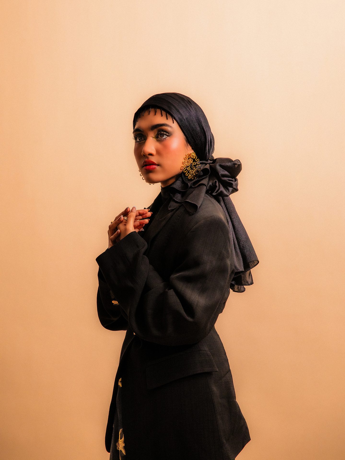 Woman in black hijab and blazer, hands clasped, looking left, with gold earrings.