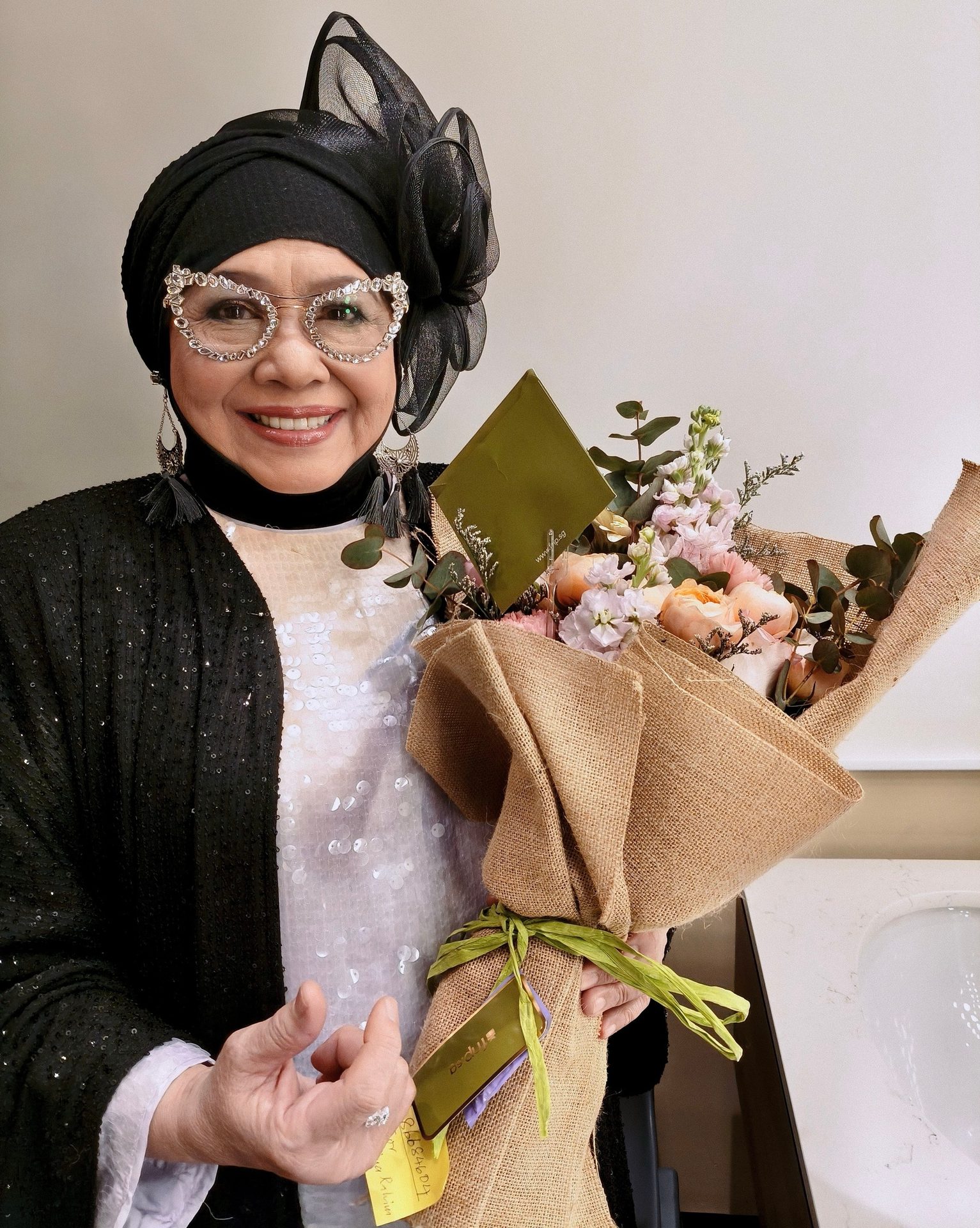Smiling woman in black headscarf and ornate glasses, holding a burlap-wrapped flower bouquet.