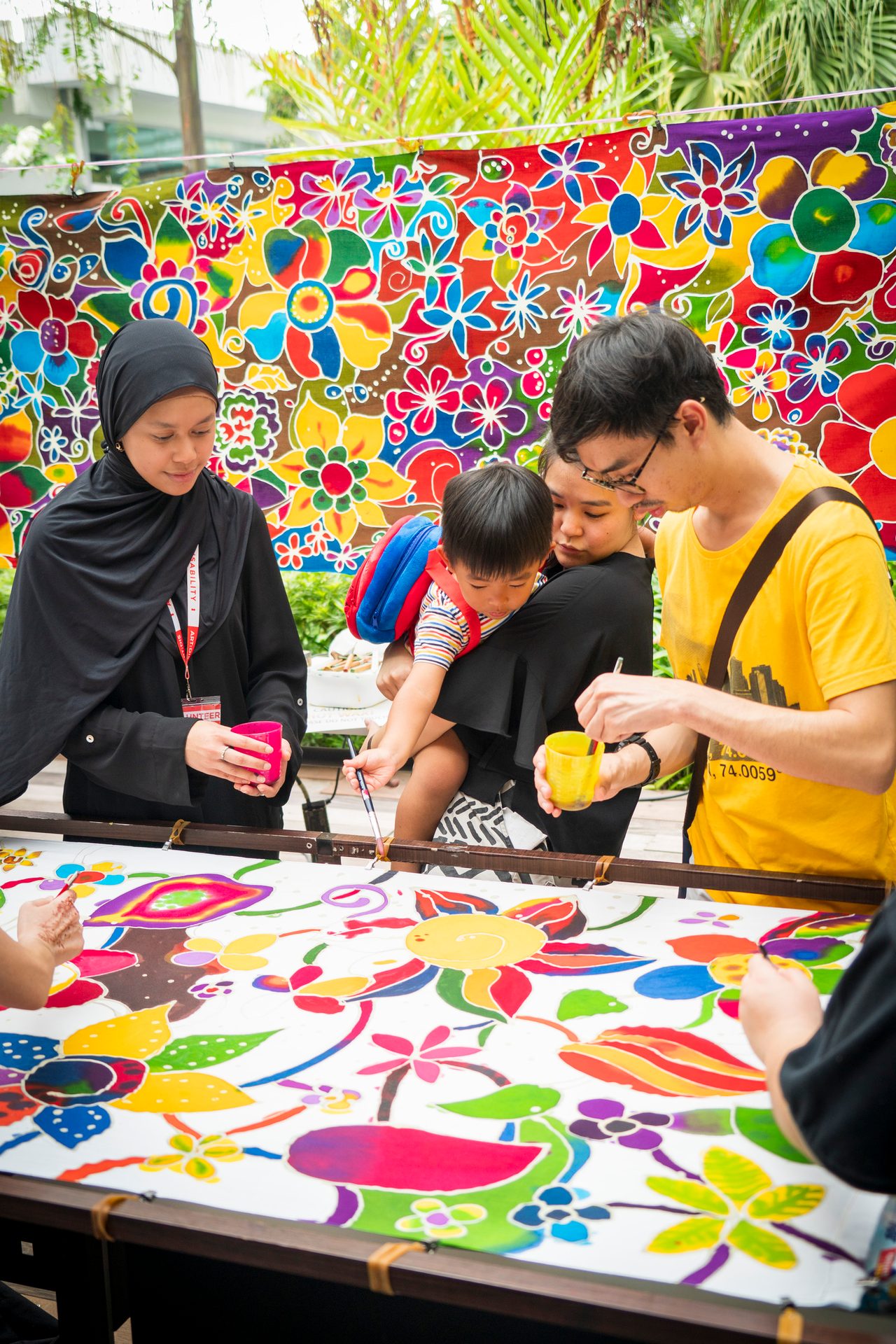 People, including a child, painting vibrant floral patterns on fabric with a colorful backdrop.
