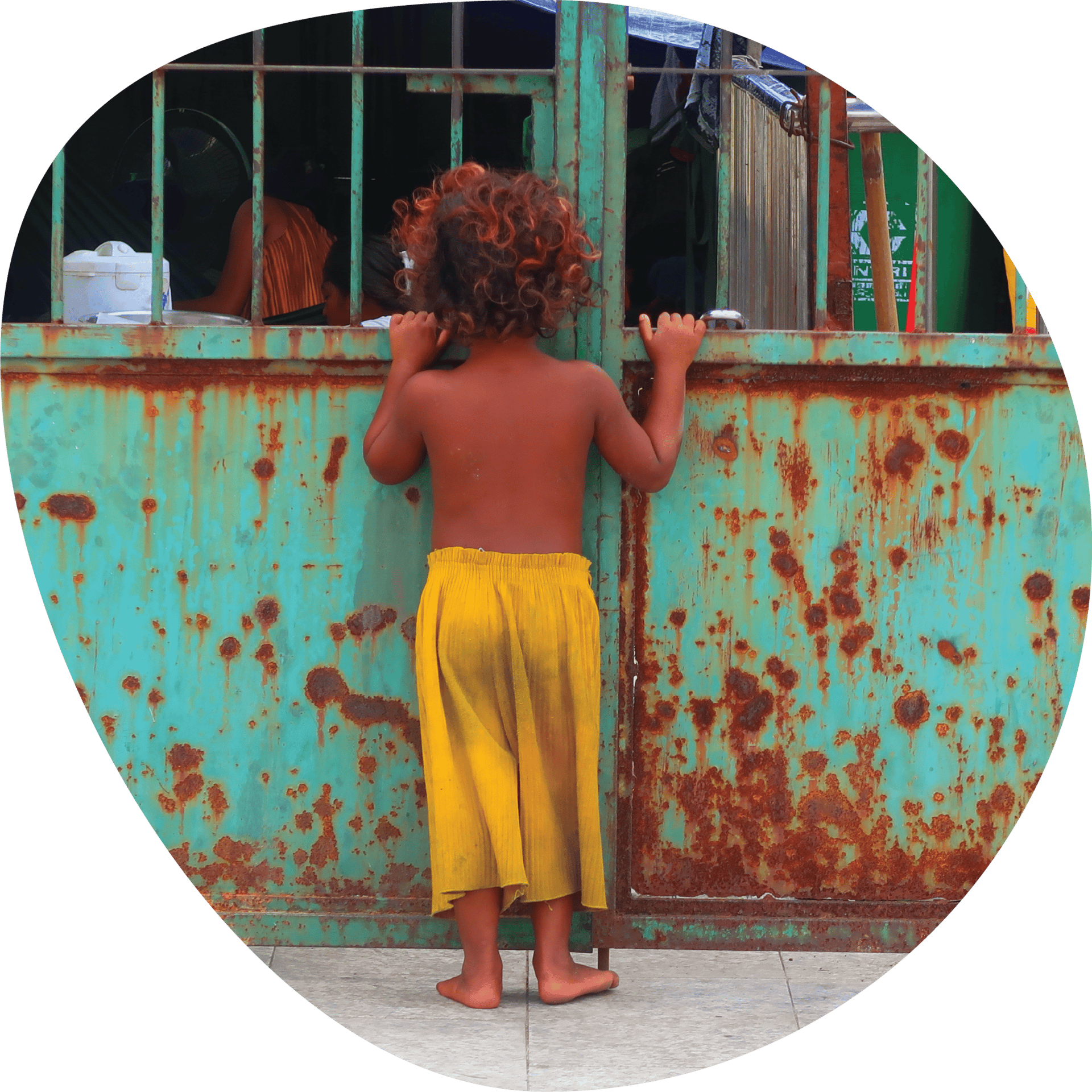 Child with red curls in yellow pants looks through a rusty green gate.