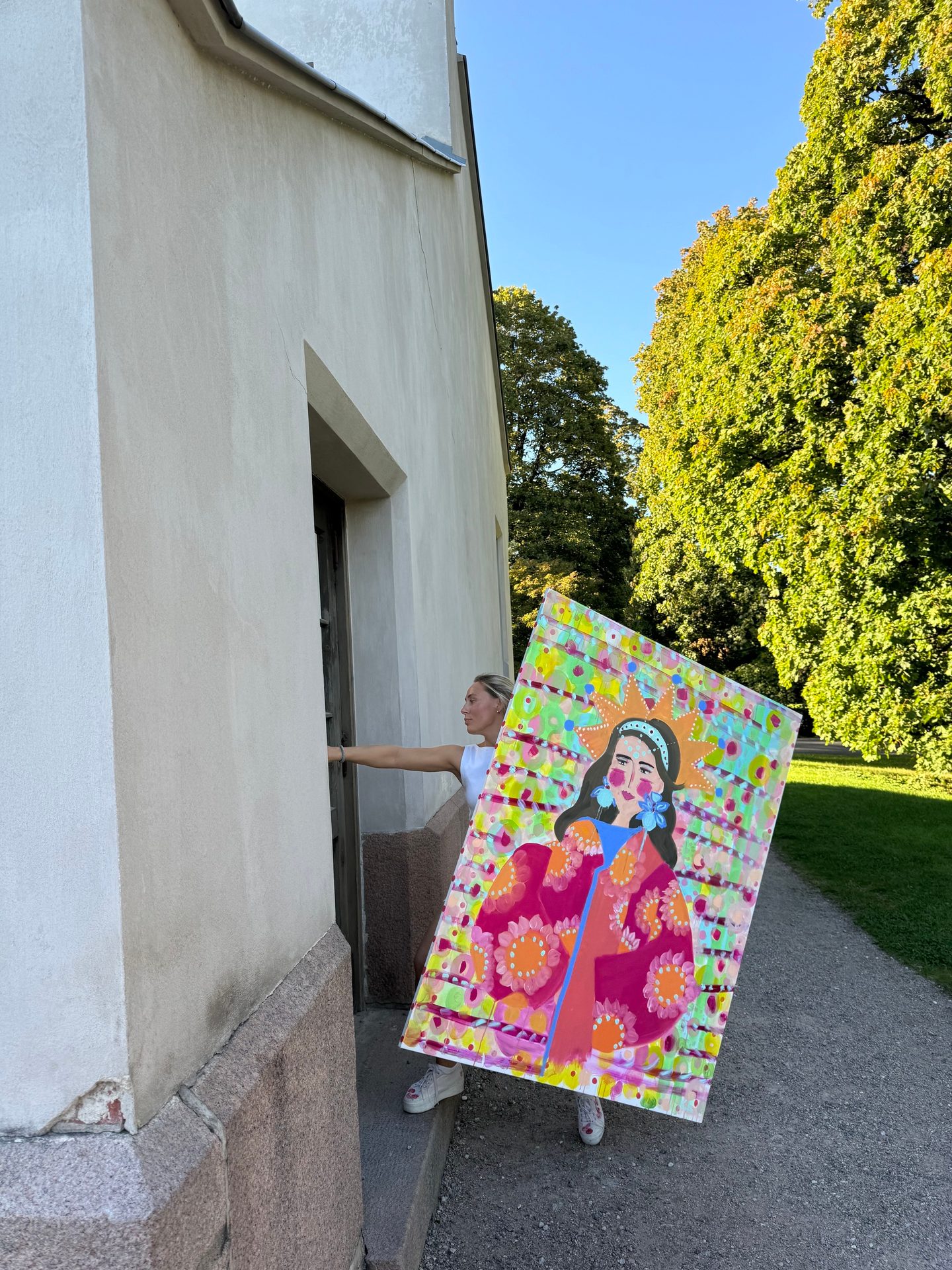 A woman holding a large, colorful painting of a woman, exiting a building onto a path.