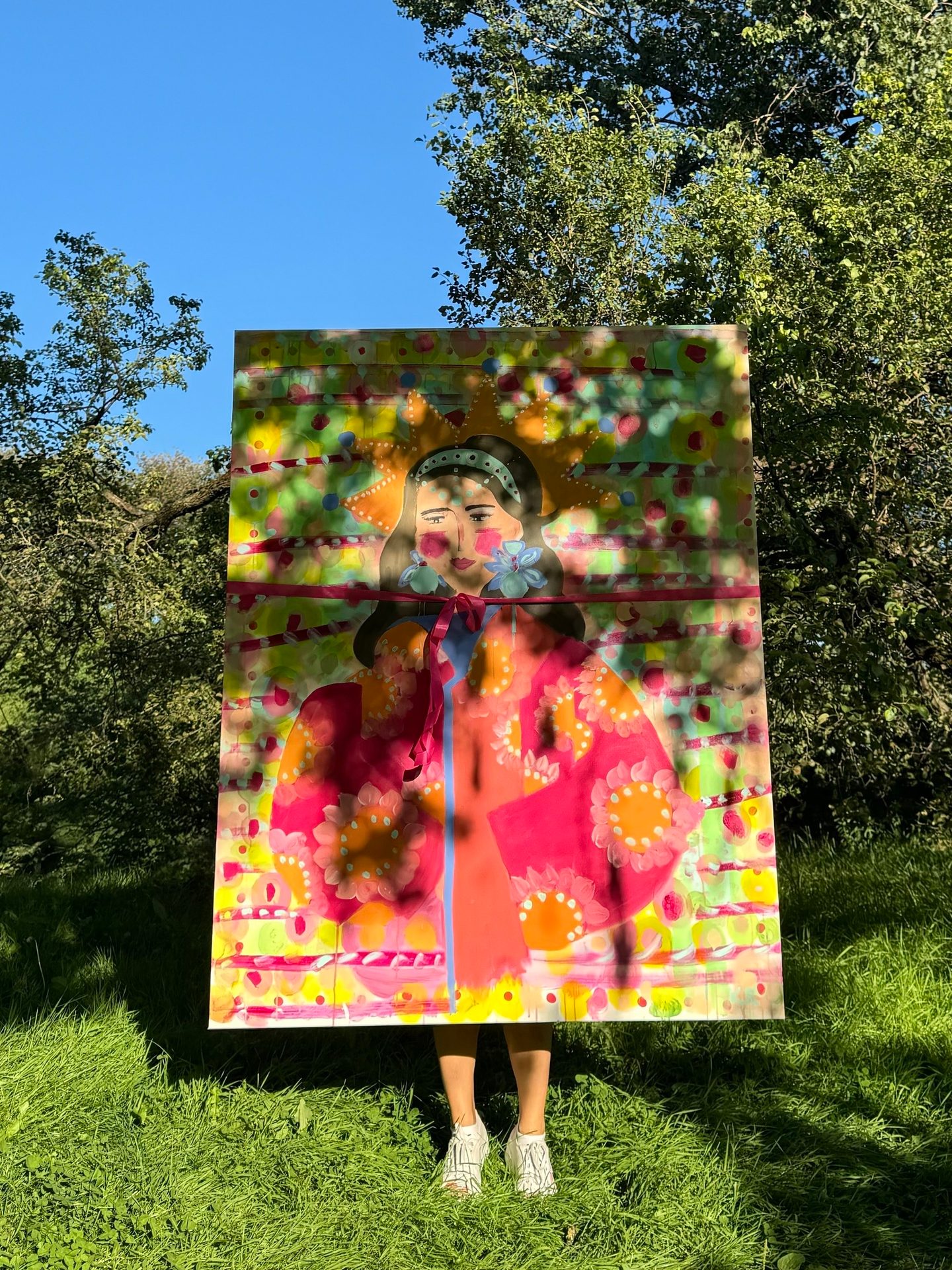 Large colorful painting of a woman held outdoors in sunlit grass with trees.